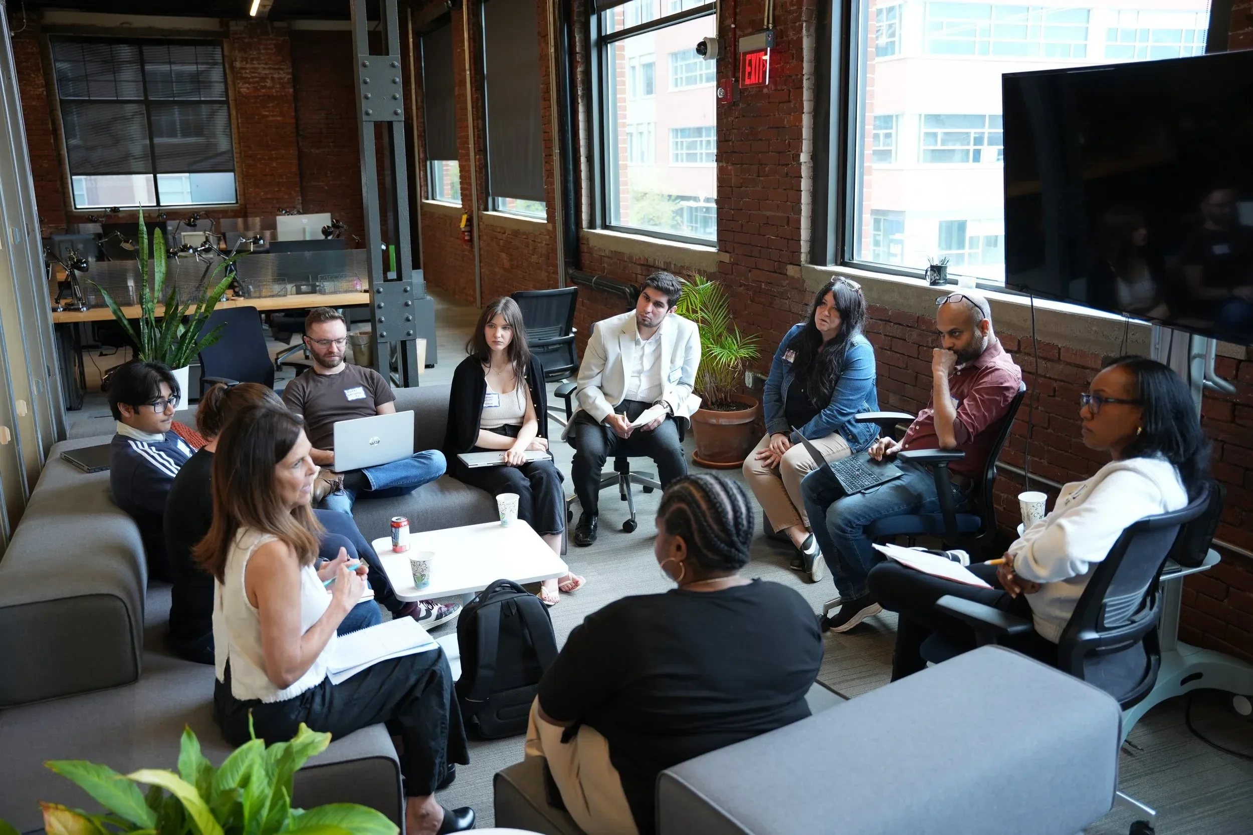 A diverse group of people sitting in a semi-circle in a modern office with exposed brick walls and large windows, participating in a meeting or discussion.