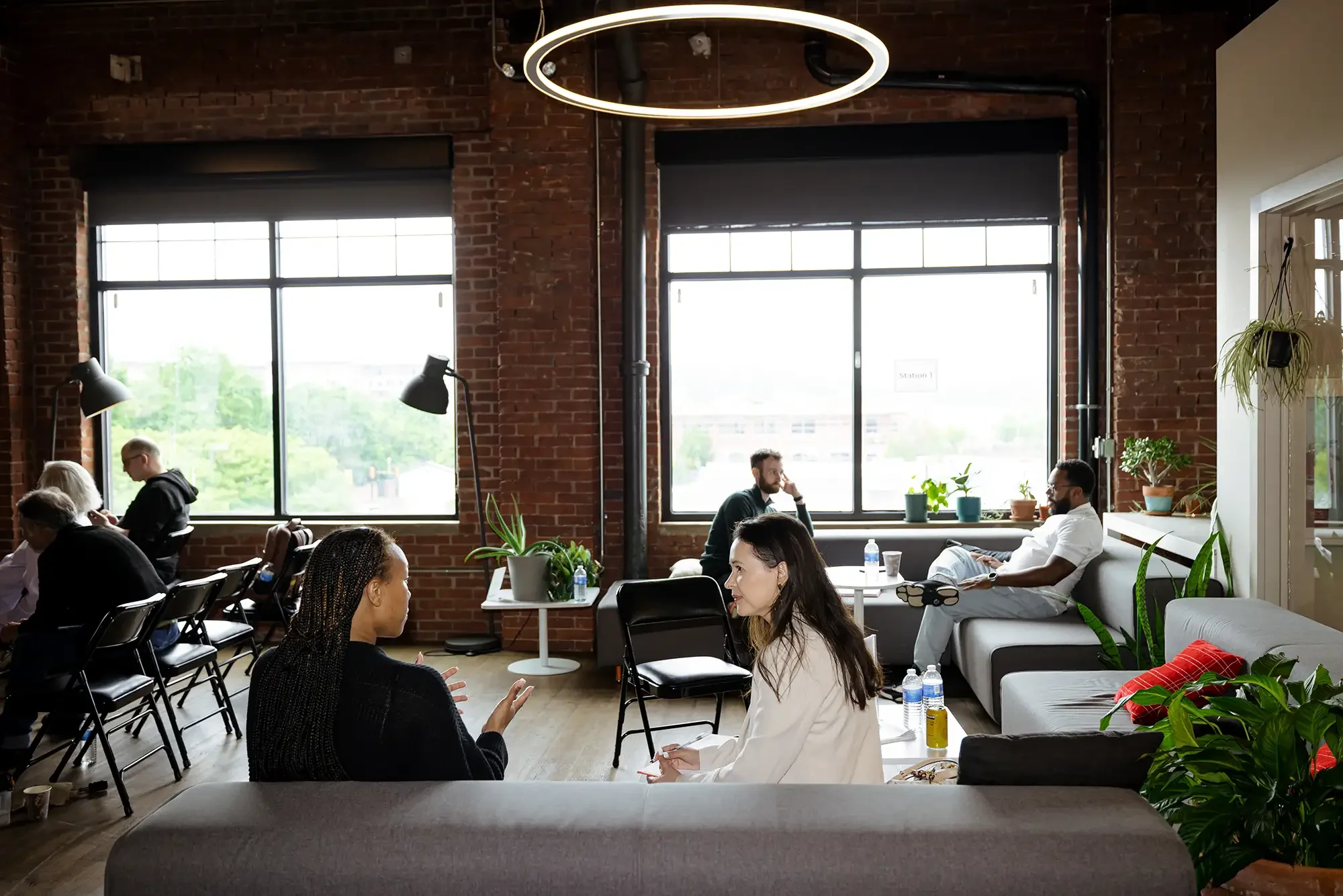 People sitting and talking in a bright industrial-style room with large windows, exposed brick walls, and indoor plants, during a meeting or event.