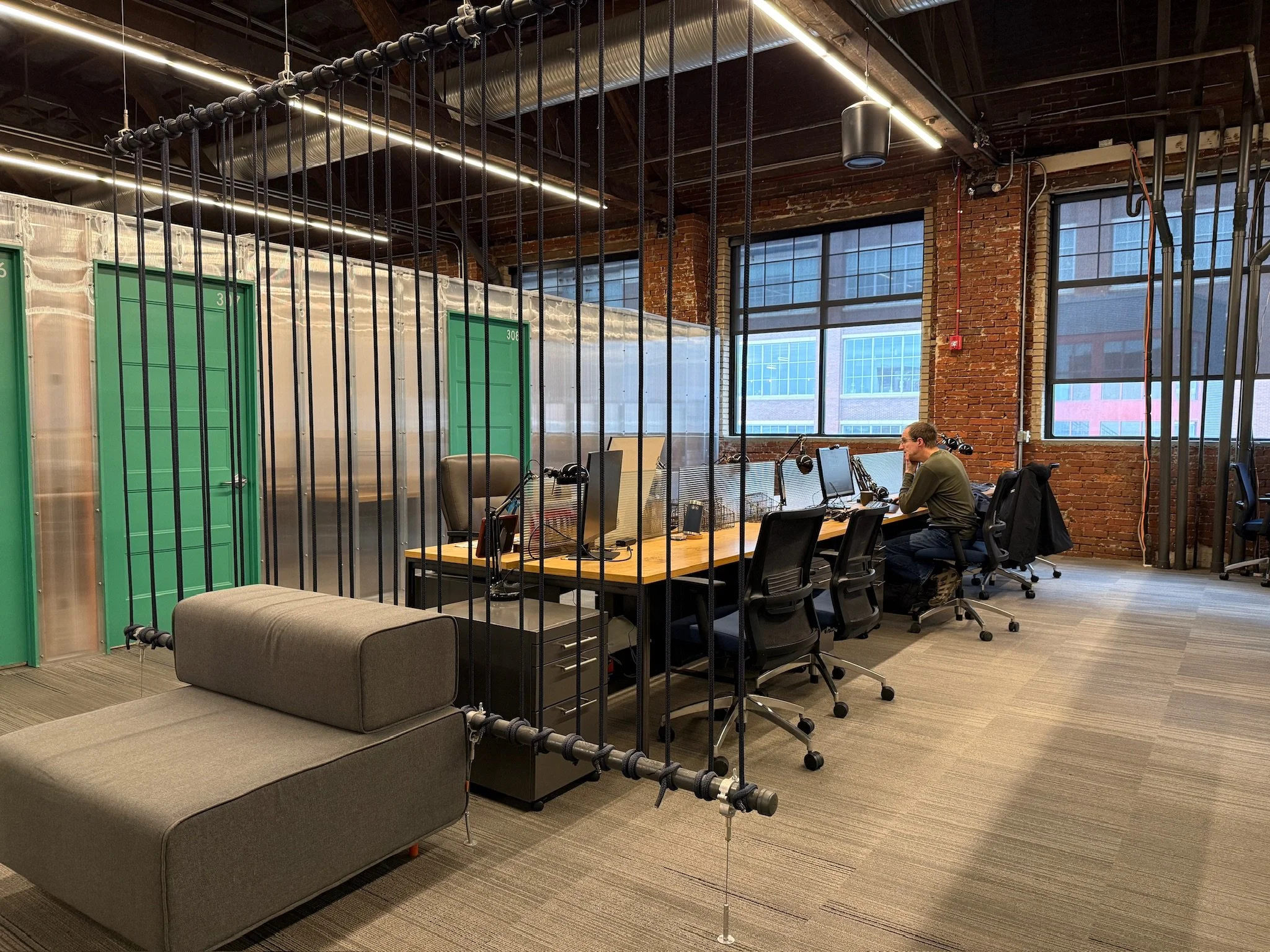 A photo of Ascender's flex desks with large windows, and a metal pipe barrier in the foreground. There are work desks with computers and chairs.