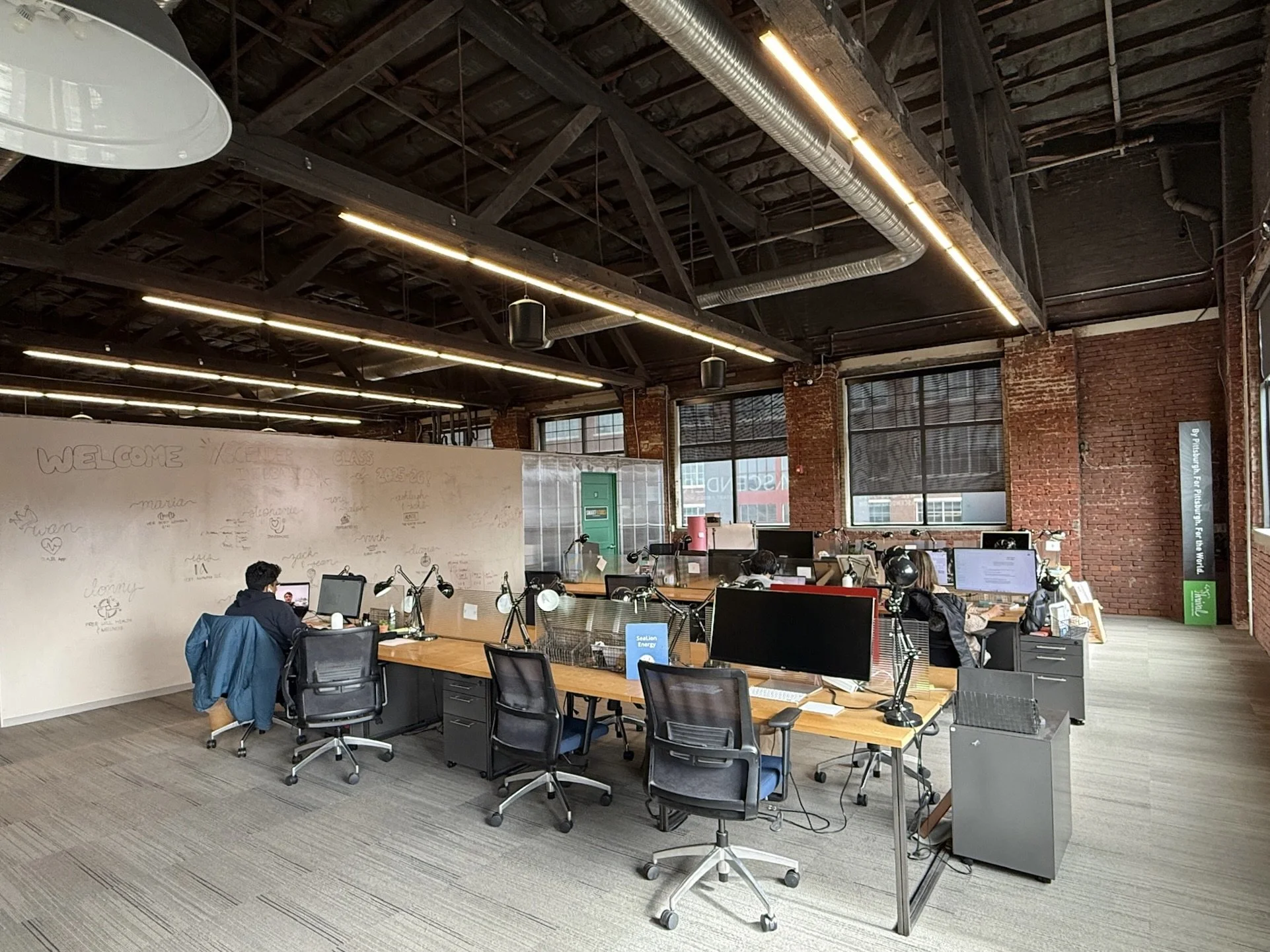 A photo of Ascender's rear dedicated desks with a high ceiling with wooden beams and ductwork, large windows, rows of desks with computers.