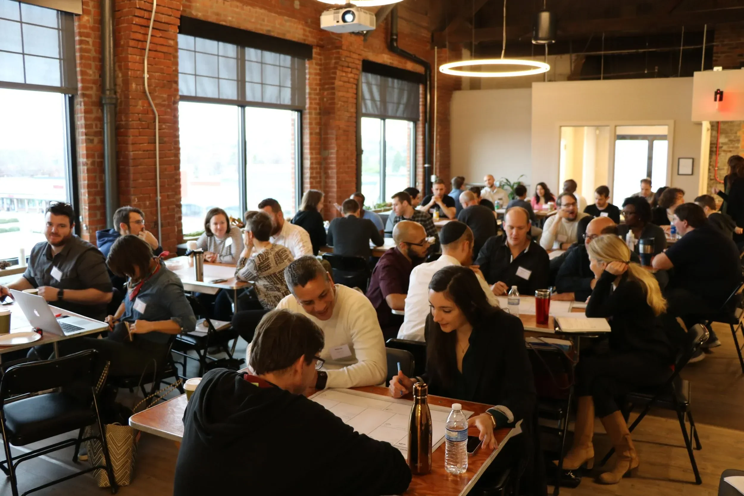A crowded indoor event in a brick-walled space with large windows, featuring many people sitting at tables engaged in discussions, some using laptops and notebooks, with bottles and water on the tables. A circular light fixture hangs from the ceiling, and a projector is mounted on the ceiling.