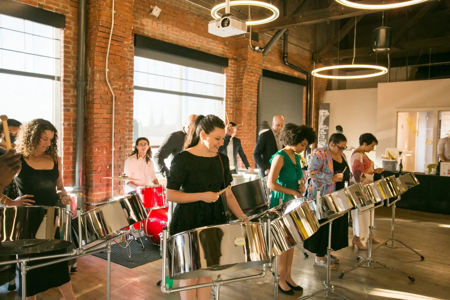 People playing steel drums in an industrial-style room with brick walls and large windows.