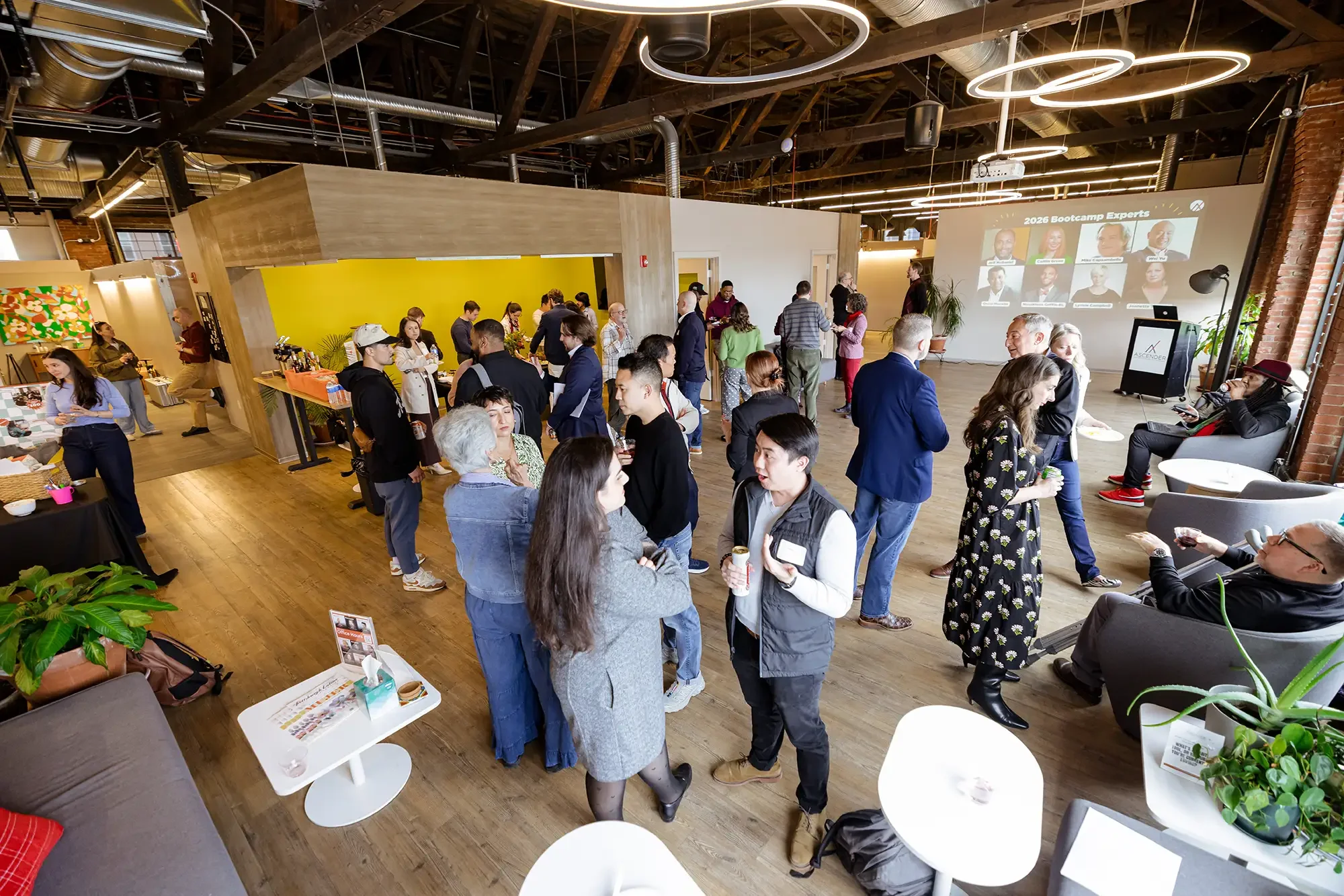 A crowded indoor event space with people socializing, some standing and some sitting. There is a large screen displaying a panel titled '2026 Bootcamp Experts' with headshots of speakers. The room has wooden floors, exposed brick and ceiling pipes, m