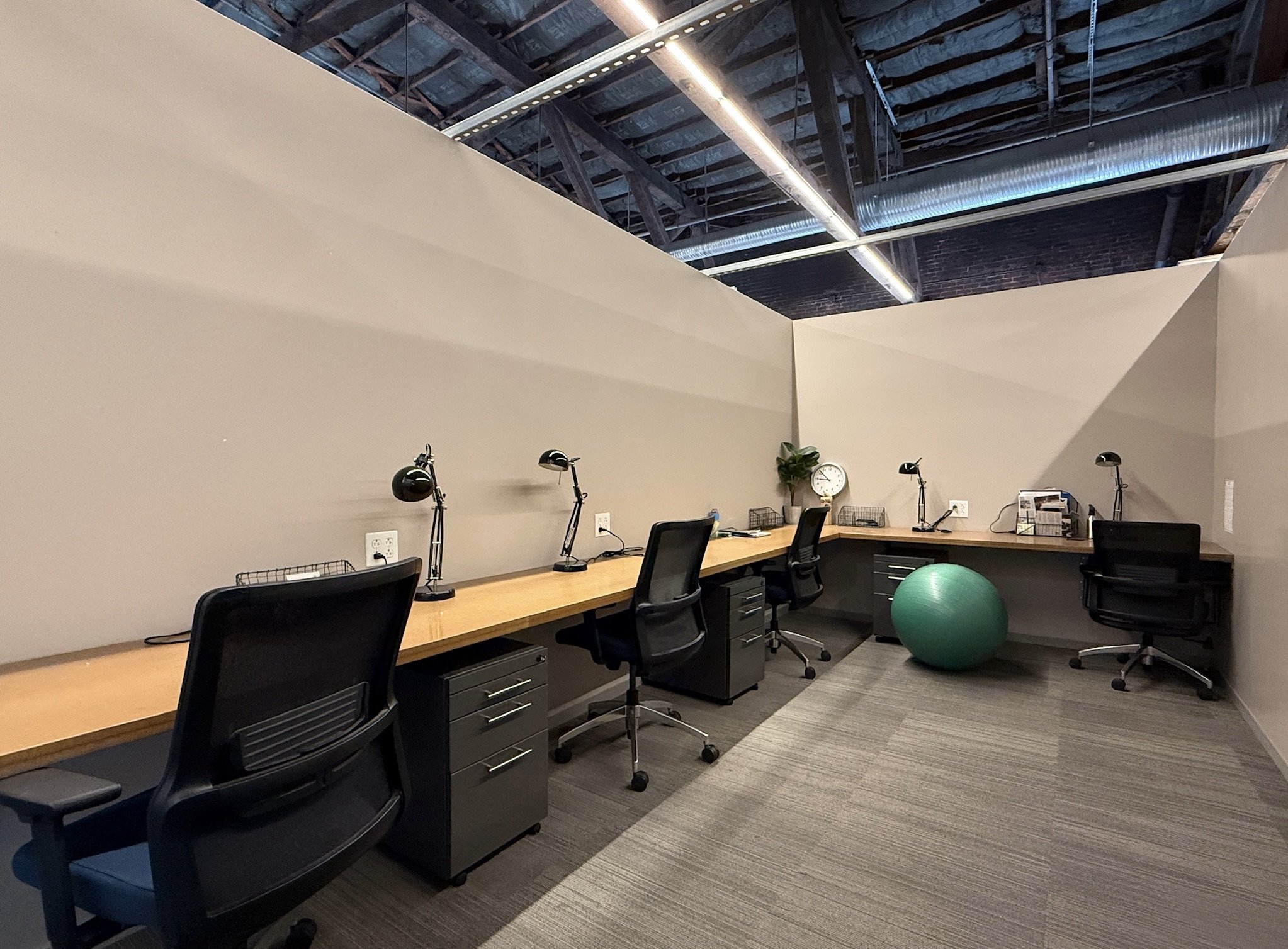 A photo of Ascender's 4-person office with a long wooden desk, black chairs, desk lamps, a clock, a green exercise ball, and some papers.
