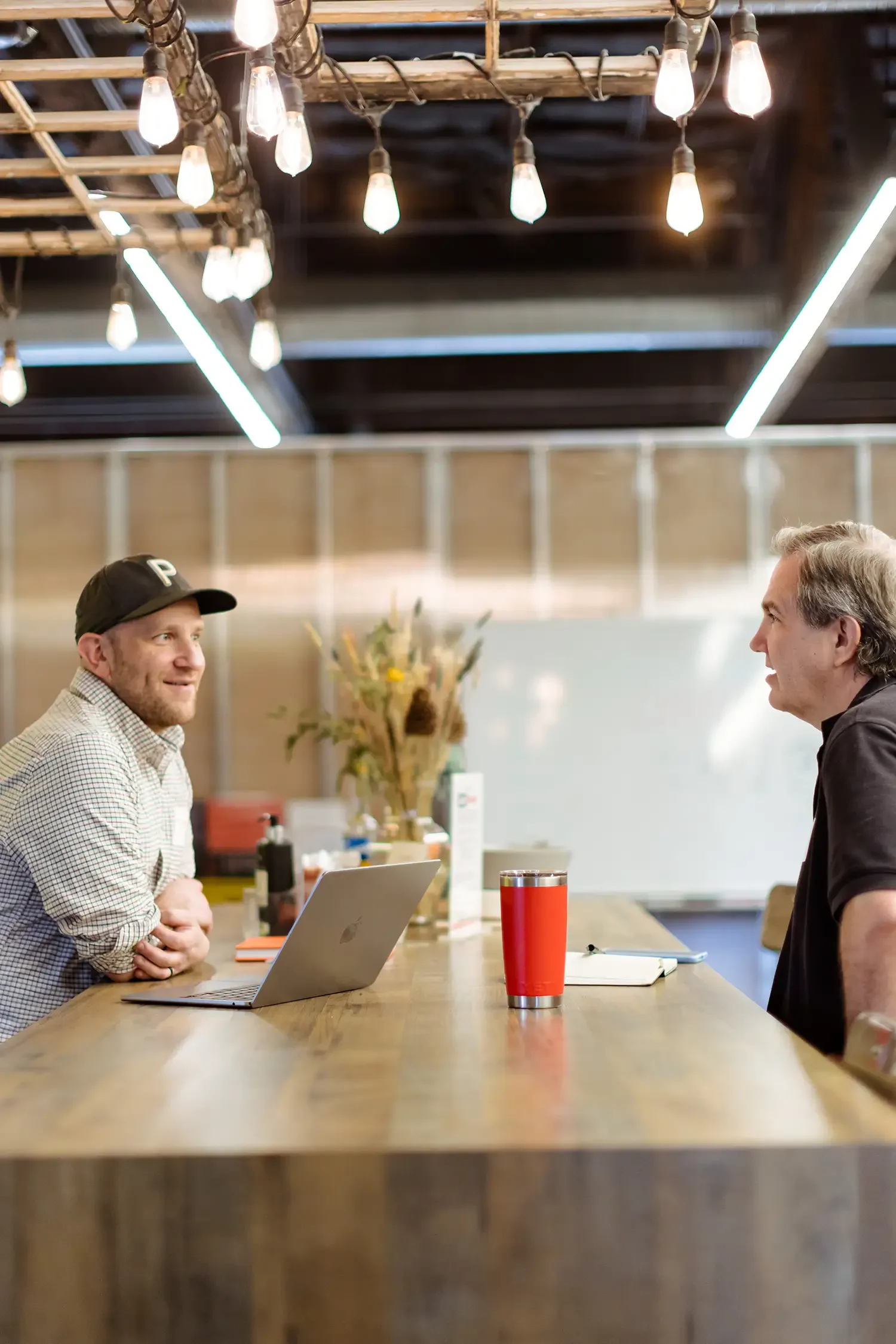 Two men are engaged in a conversation across a long wooden table in a modern office or coworking space. One man wears a baseball cap labeled 'P' and a checkered shirt, sitting with a laptop in front of him. The other man, with gray hair, wears a dark