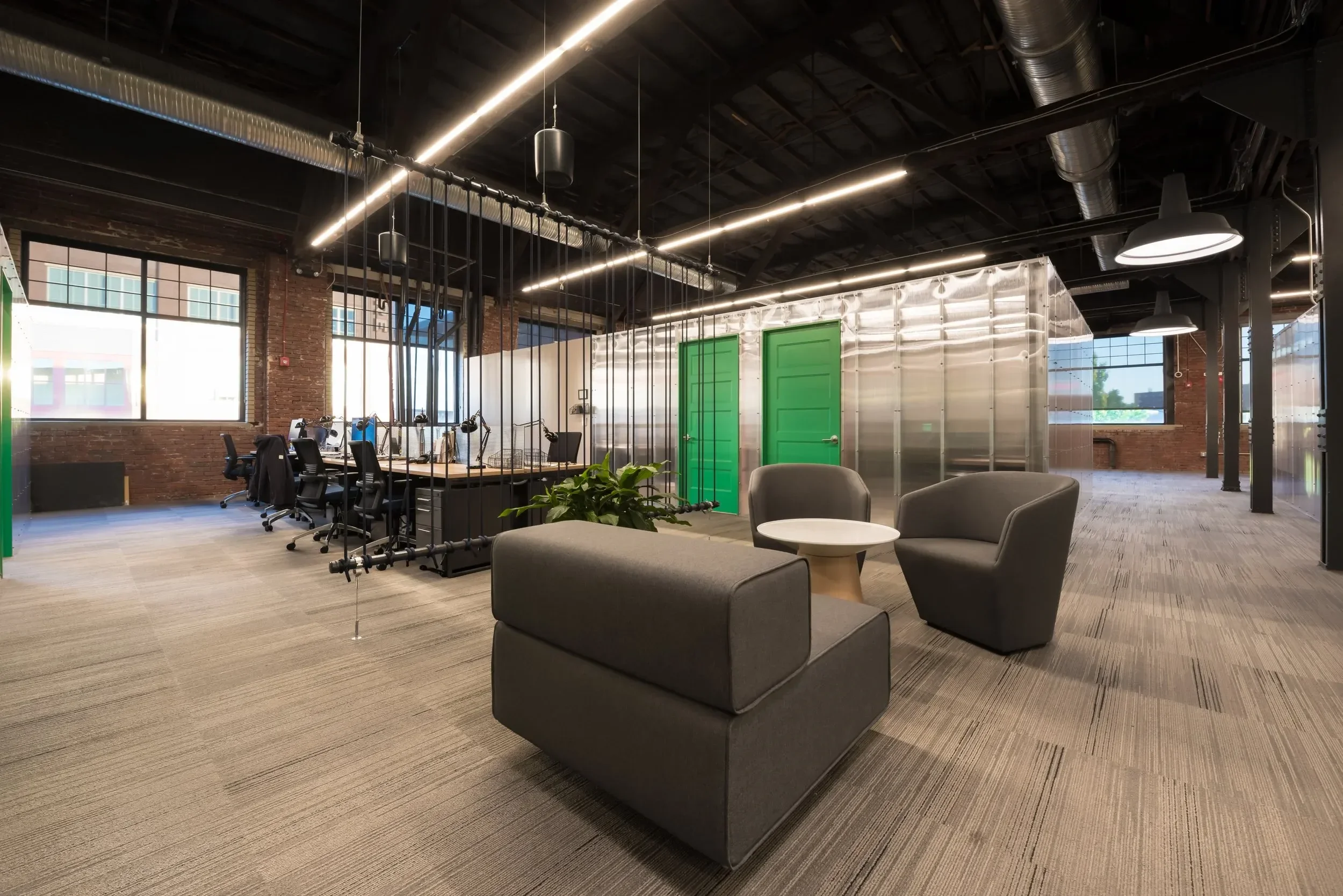 A photo of Ascender's flex desks. There are multiple workstations with black chairs, a lounge area with gray sofas, and green doors. Overhead lighting and a small indoor plant are also visible.
