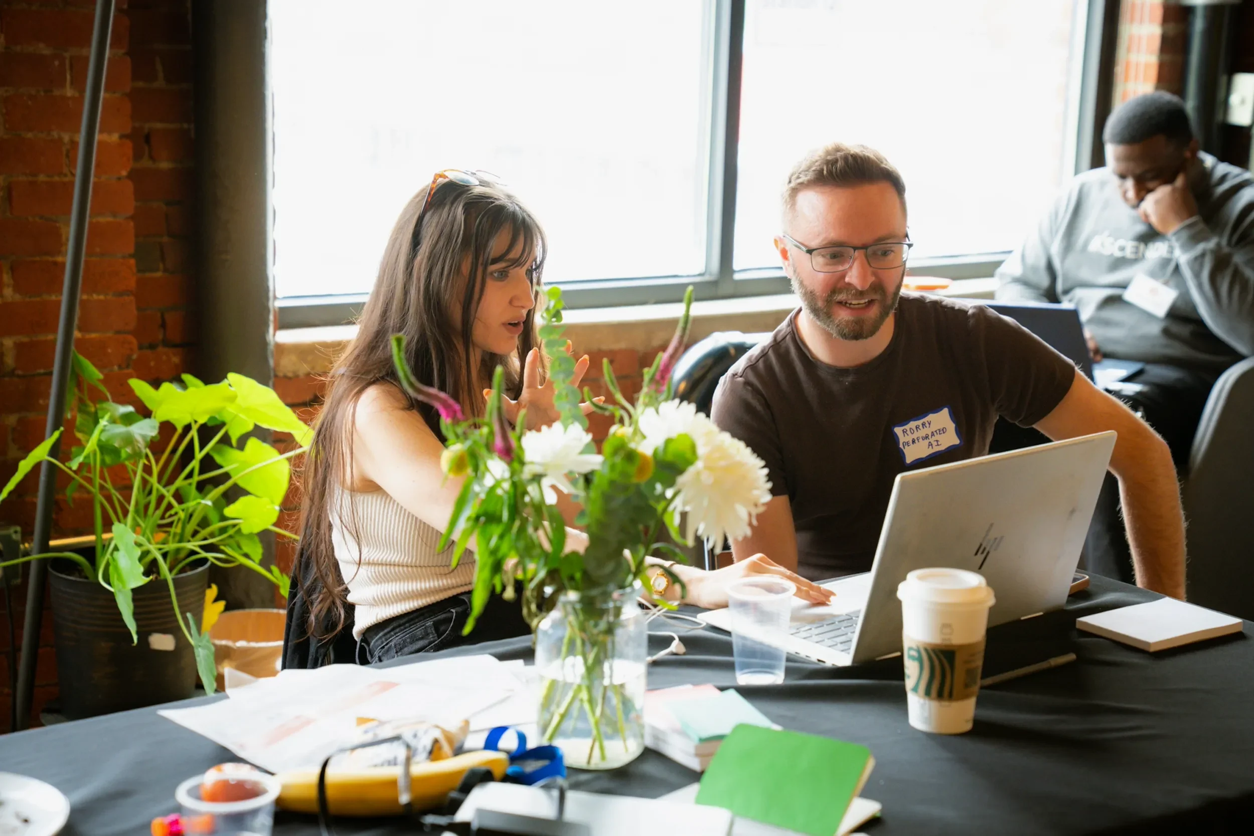 Two people seated at a table working on laptops, with plants and flowers in the foreground, near a large window with brick wall behind.