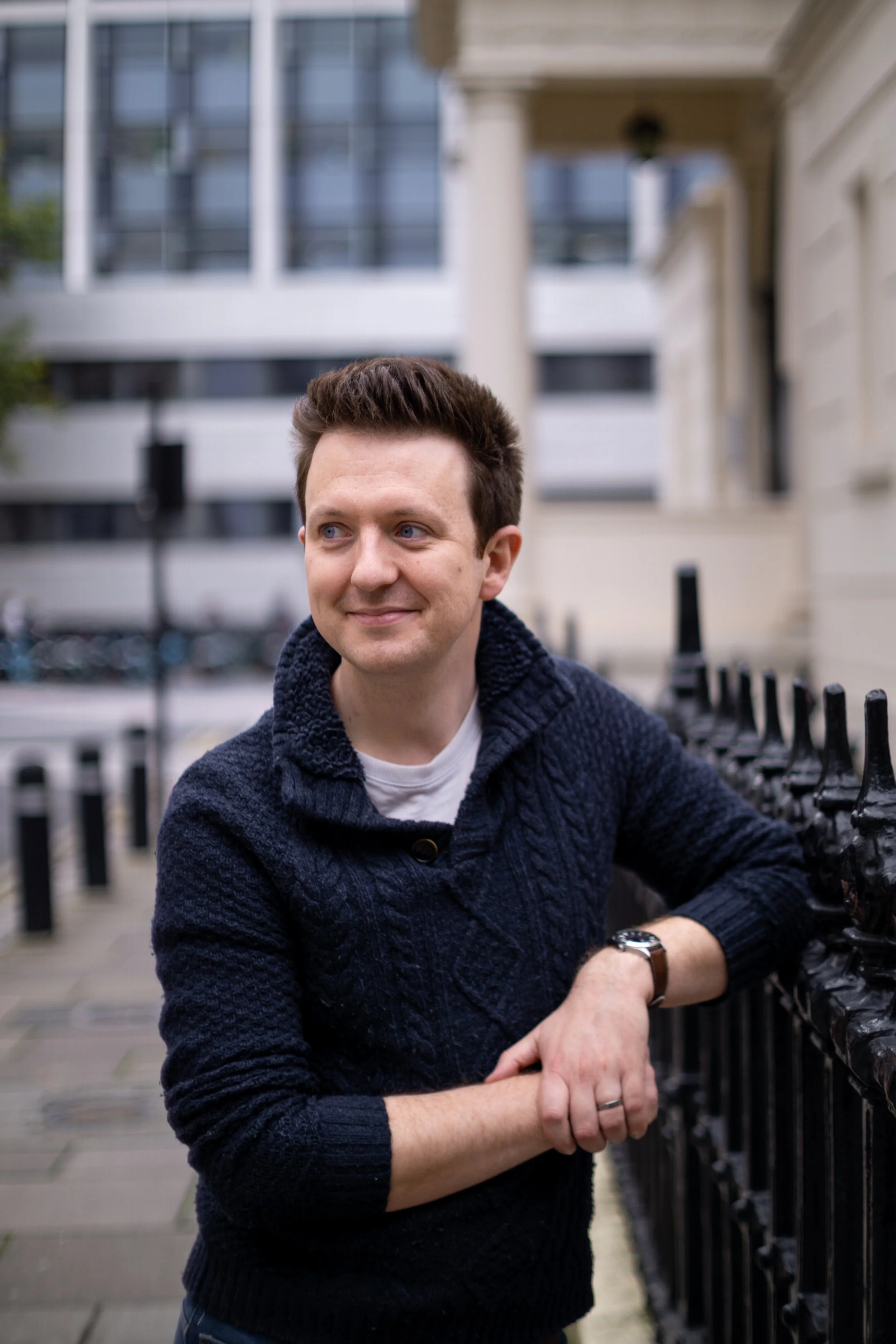 A smiling man with brown hair, wearing a dark blue sweater, leaning on a black fence outdoors in an urban setting.