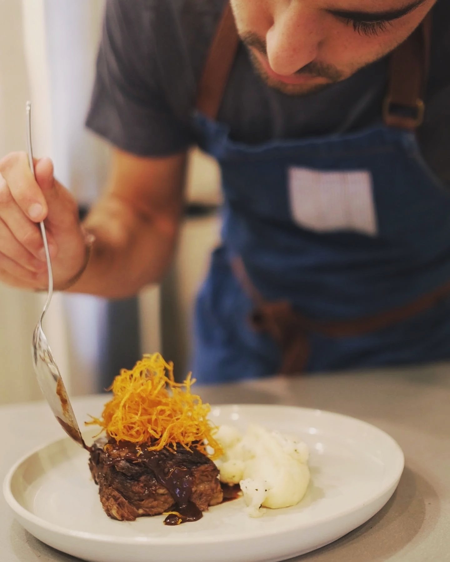 A chef or cook is carefully plating a steak topped with shredded cheddar cheese and dark gravy, accompanied by mashed potatoes on a white plate.