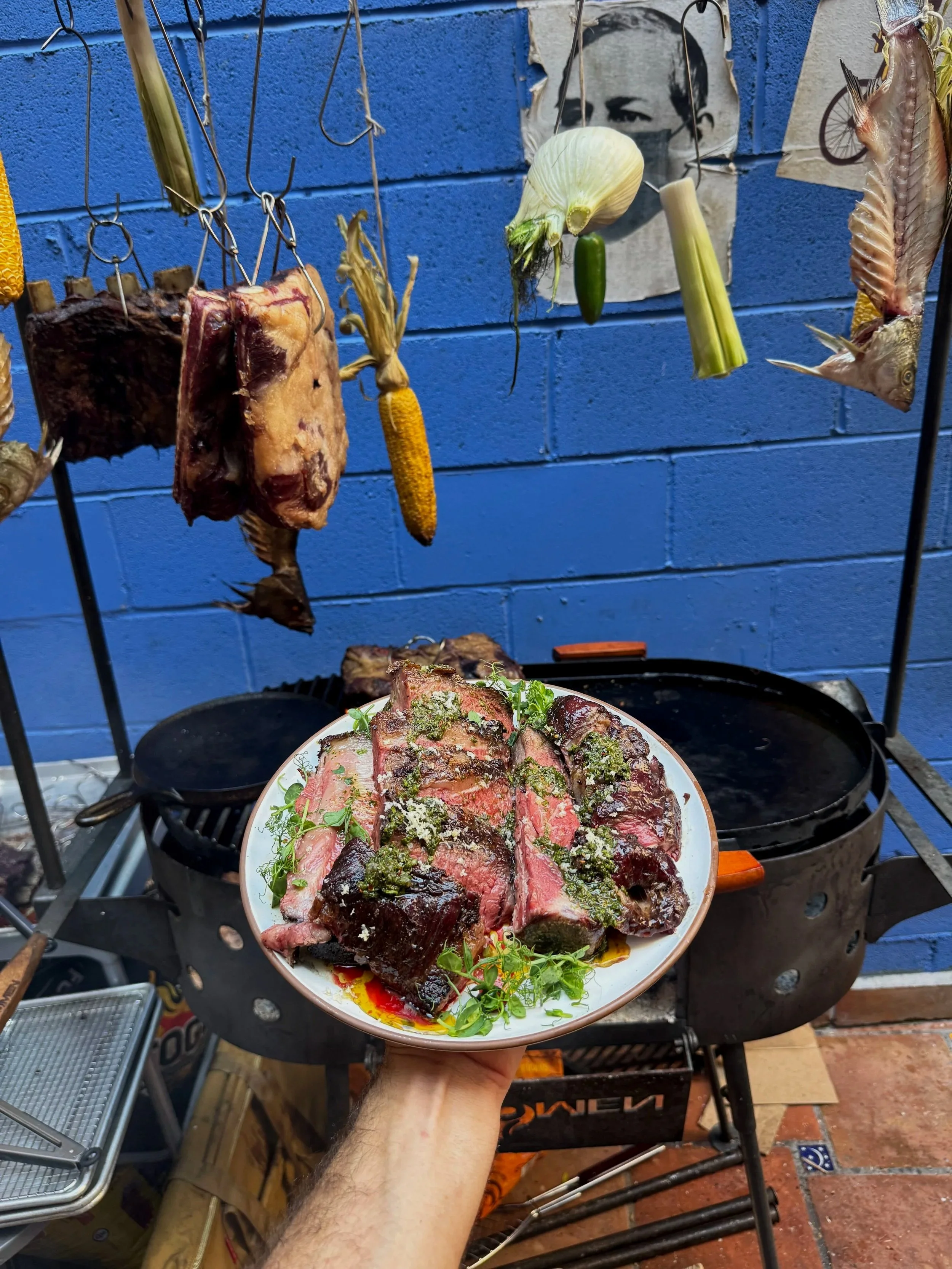A plate of cooked steak garnished with herbs and sauce, held in front of a barbecue grill with hanging meats, vegetables, and seafood, set against a blue brick wall.