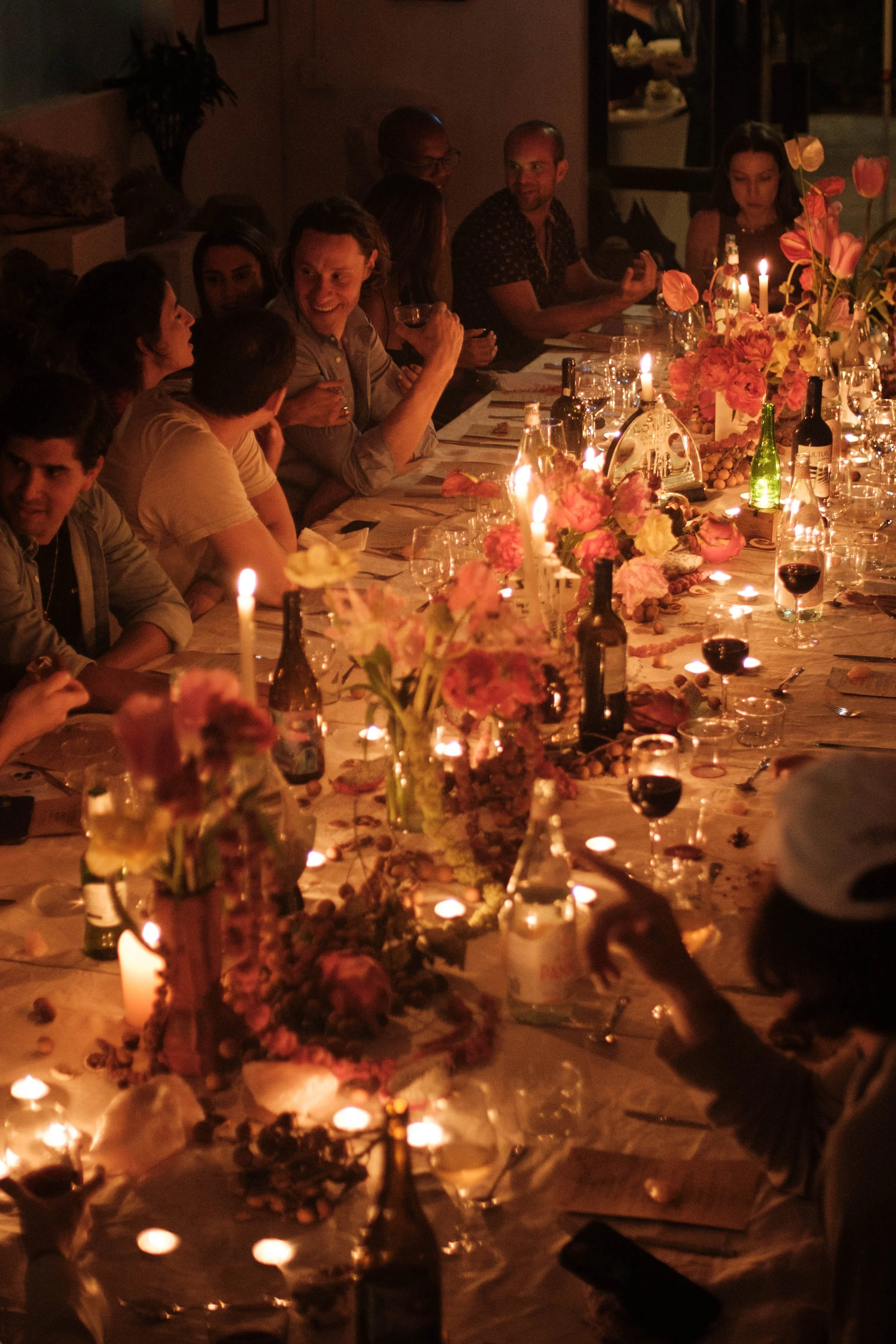 People sitting at a long dinner table decorated with flowers, candles, and bottles, enjoying a gathering in a warmly lit environment.