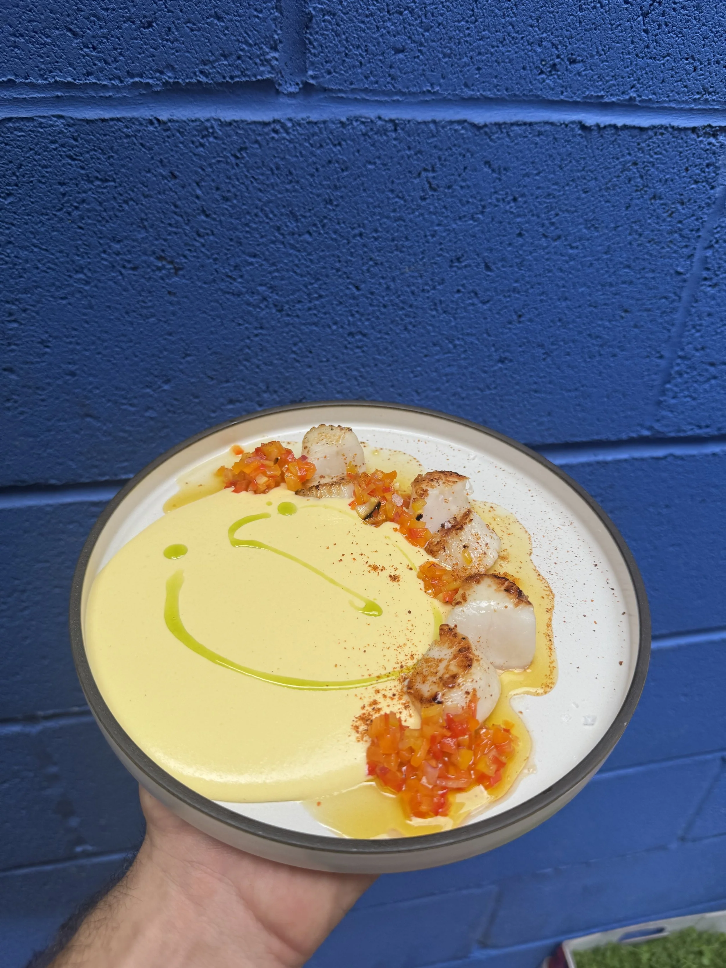 A bowl of yellow soup garnished with chopped vegetables, grilled scallops, a drizzle of green oil, and a broad smiley face design made with oil on the soup's surface, held in front of a blue brick wall.