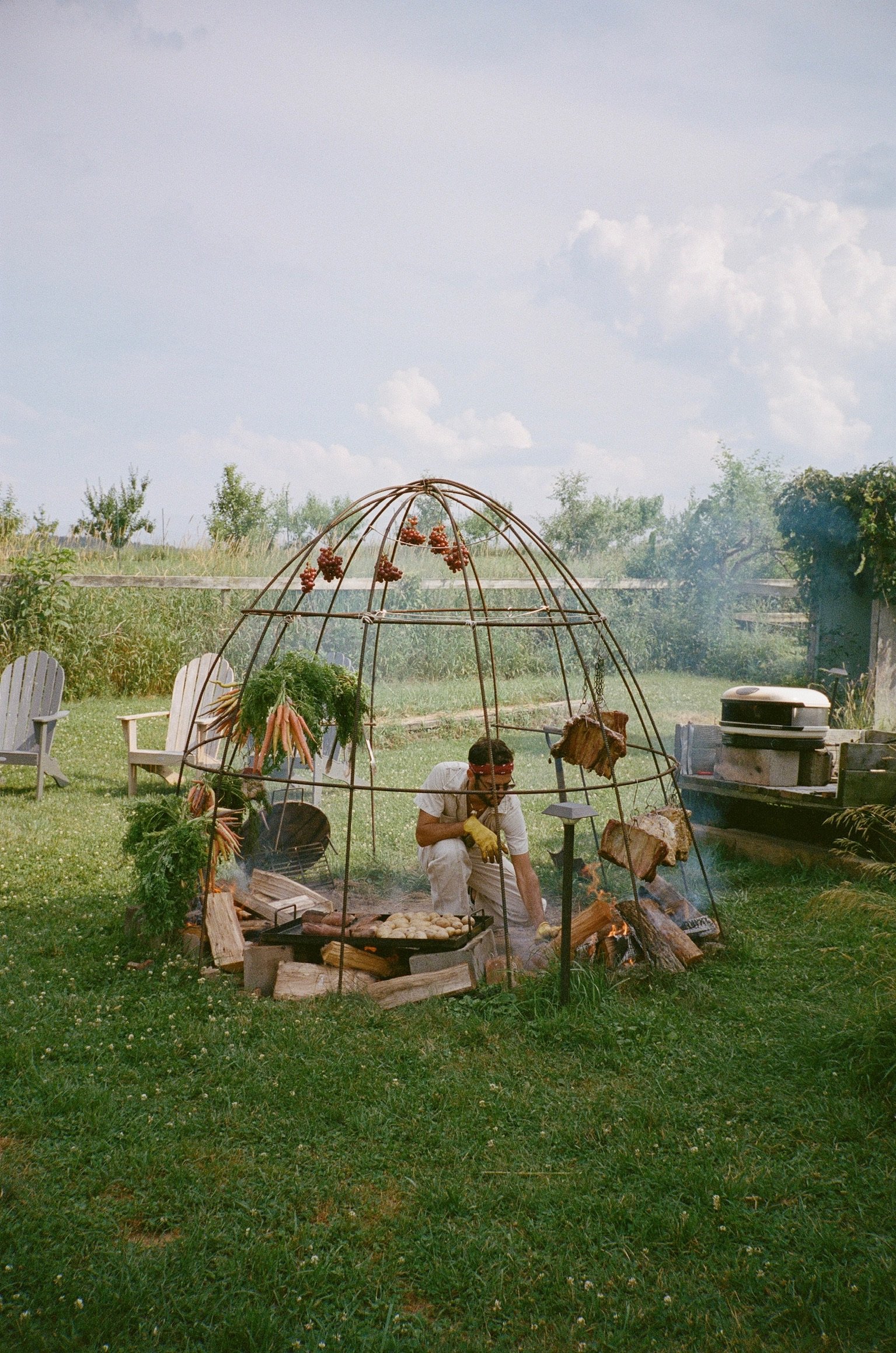 Man cooking food on open fire inside a metal wire dome structure outdoors, with greenery and sky in background.