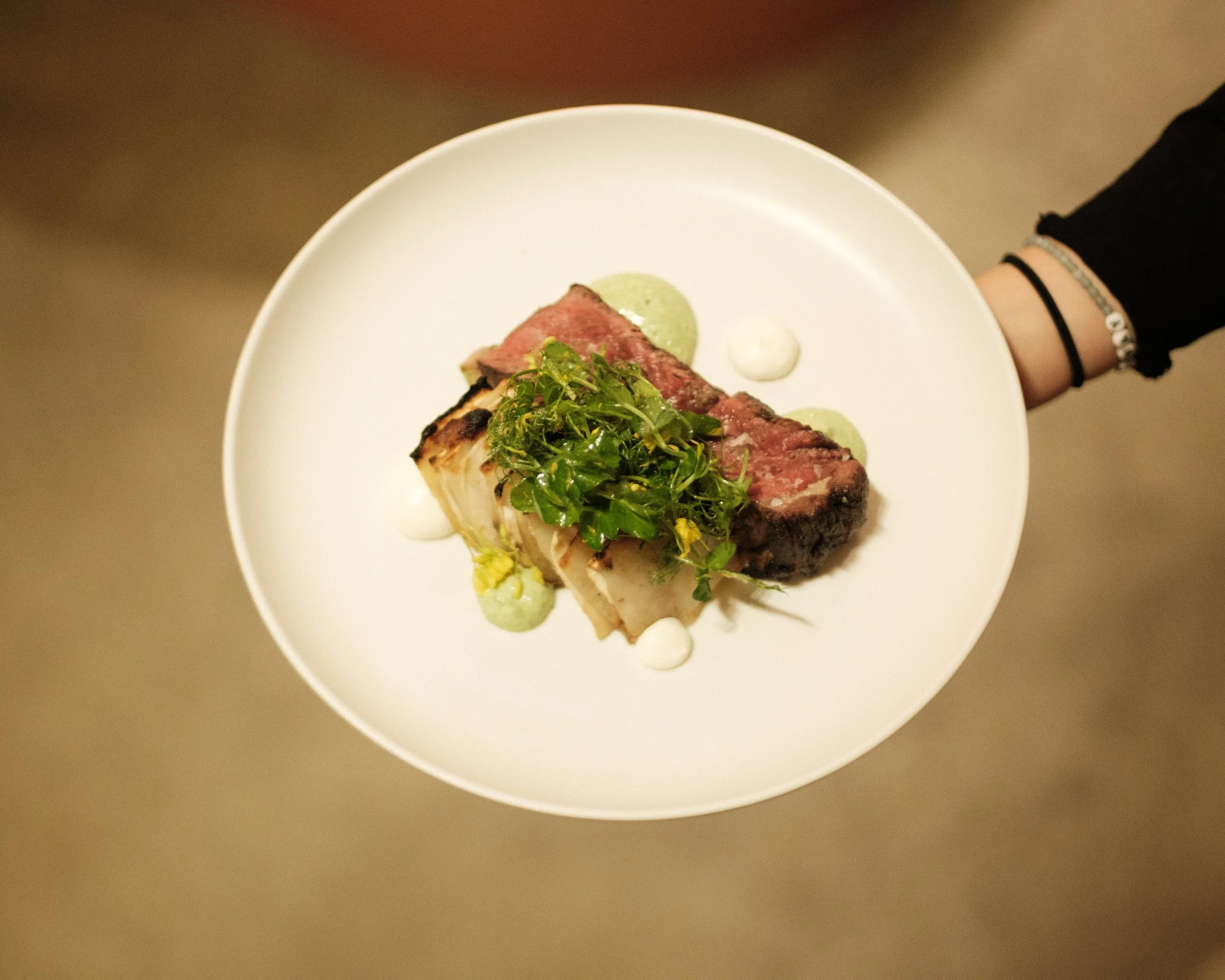 A white plate with a serving of cooked meat topped with greens, accompanied by dollops of white and green sauces, held by a person with black long-sleeve shirt and bracelets.