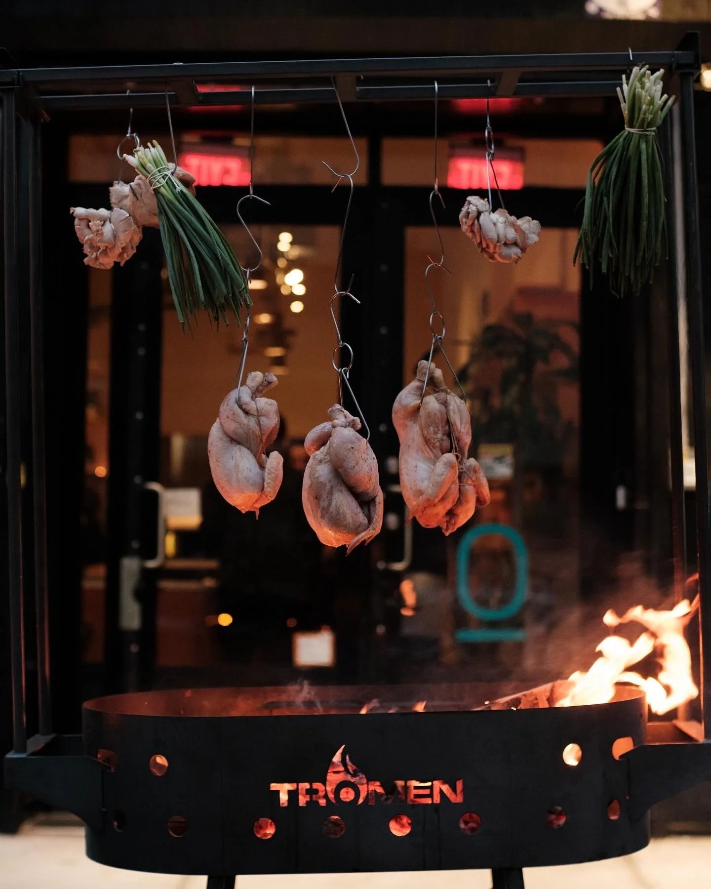 Chicken being roasted over an open flame on a vertical rotisserie with herbs and vegetables hanging above.