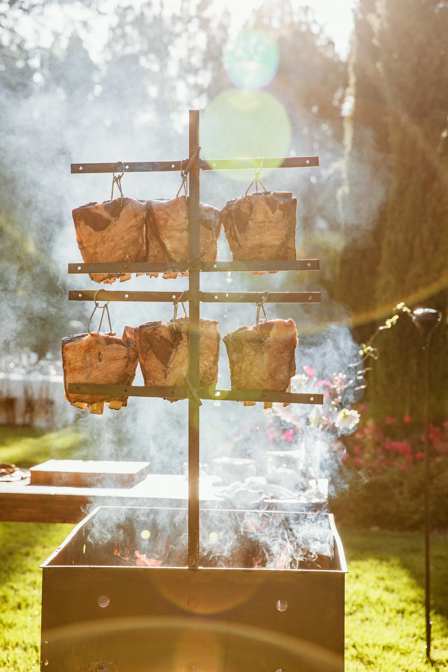 Chunks of meat being grilled on a vertical rotisserie over an open flame in an outdoor setting, sunlight filtering through trees in the background.