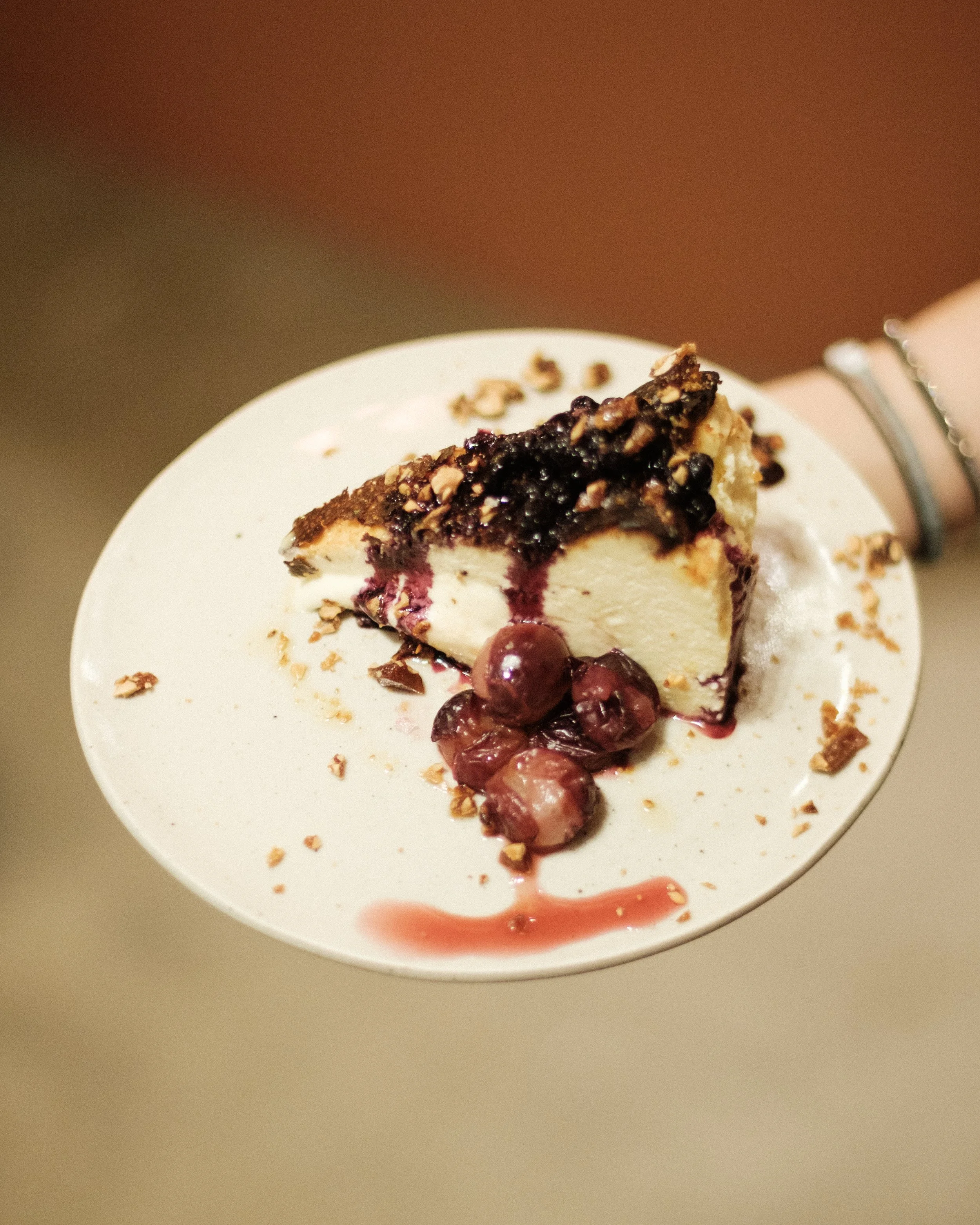 A slice of cheesecake topped with blueberry compote and chopped nuts, with cherries and cherry syrup on a white plate.