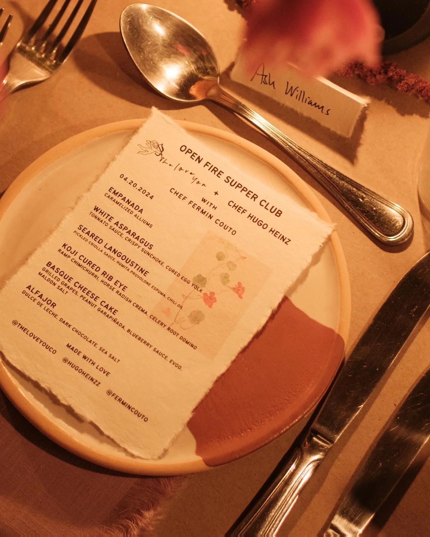 A restaurant menu on a plate with a torn edge, listing dishes such as empanada, white asparagus, seared langoustine, Kobe cured rib eye, basque cheese cake, and alfajor, with a floral pattern and handwritten notes. A silver spoon and fork are placed beside the menu, and a name tag that reads "Ash Williams" is nearby.