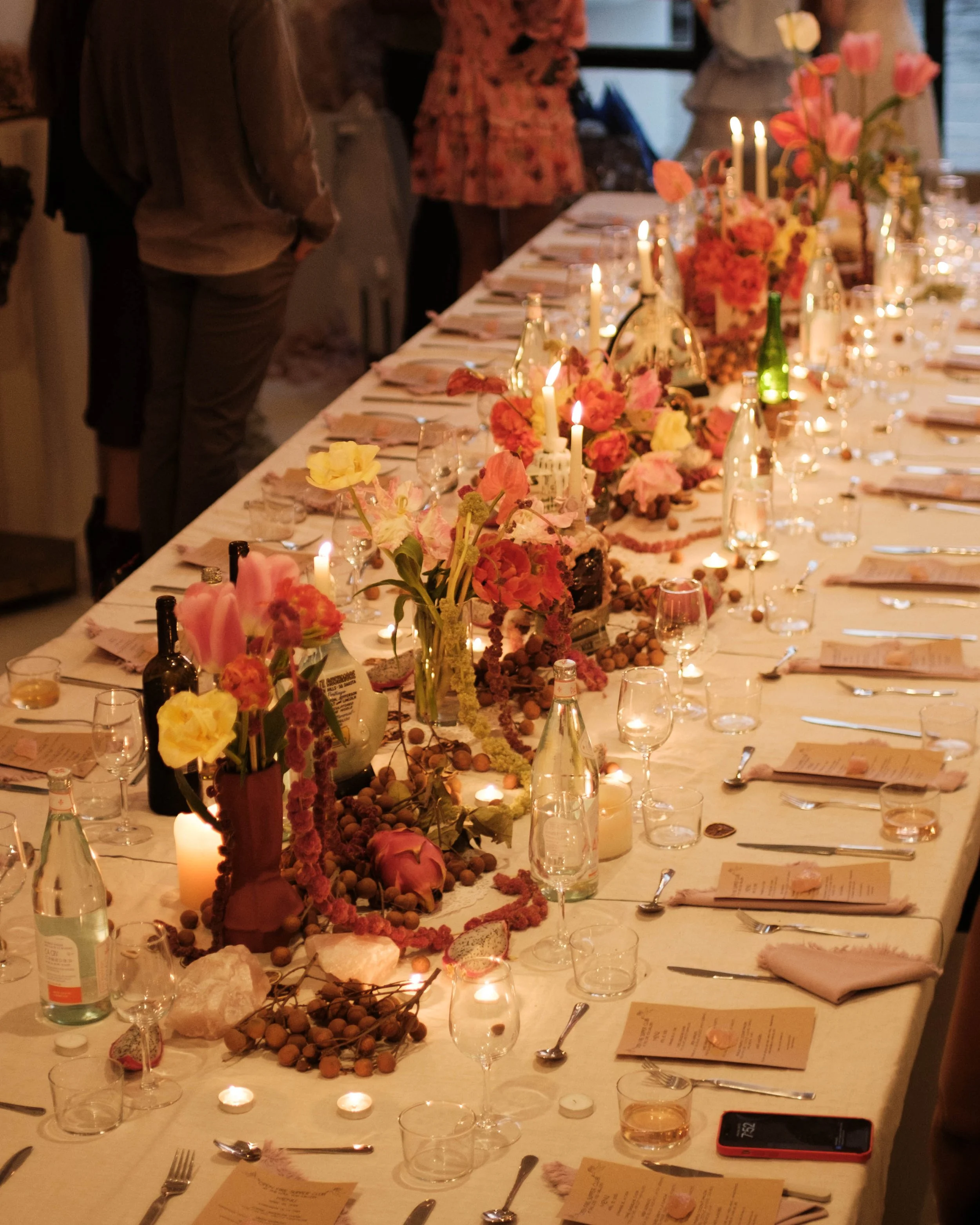 Long banquet table decorated with floral arrangements, candles, bottles, and glassware at a dinner party or celebration