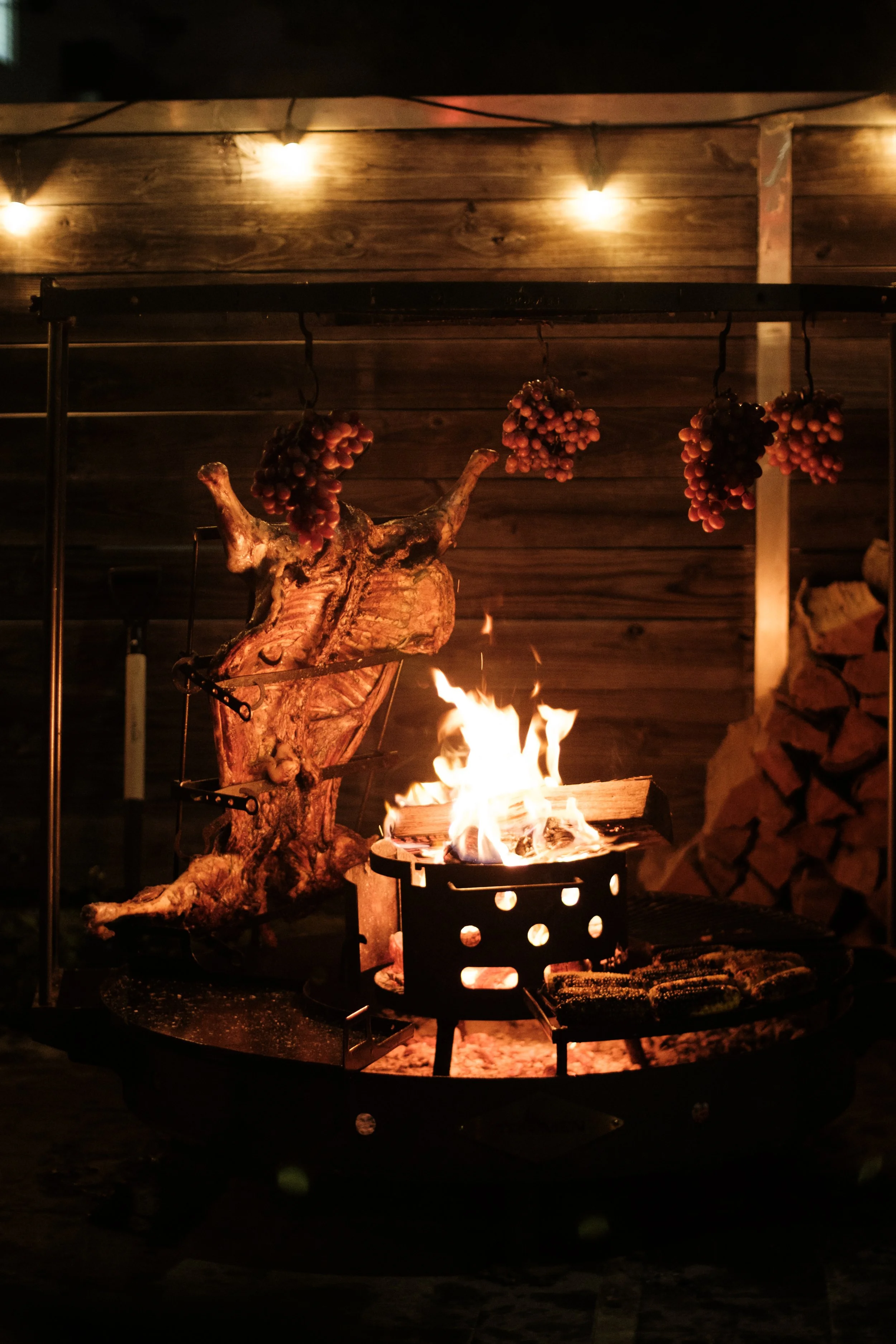 A fire pit with a large piece of firewood, grape clusters hanging above, and wood stacked to the right, with string lights overhead in a dark outdoor setting.