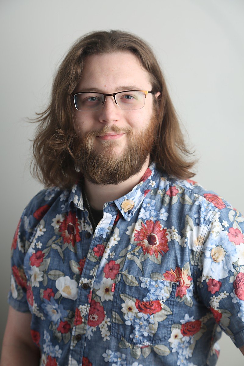Portrait of a young man with shoulder-length brown hair, beard, glasses, in a floral shirt, standing against a plain light background.