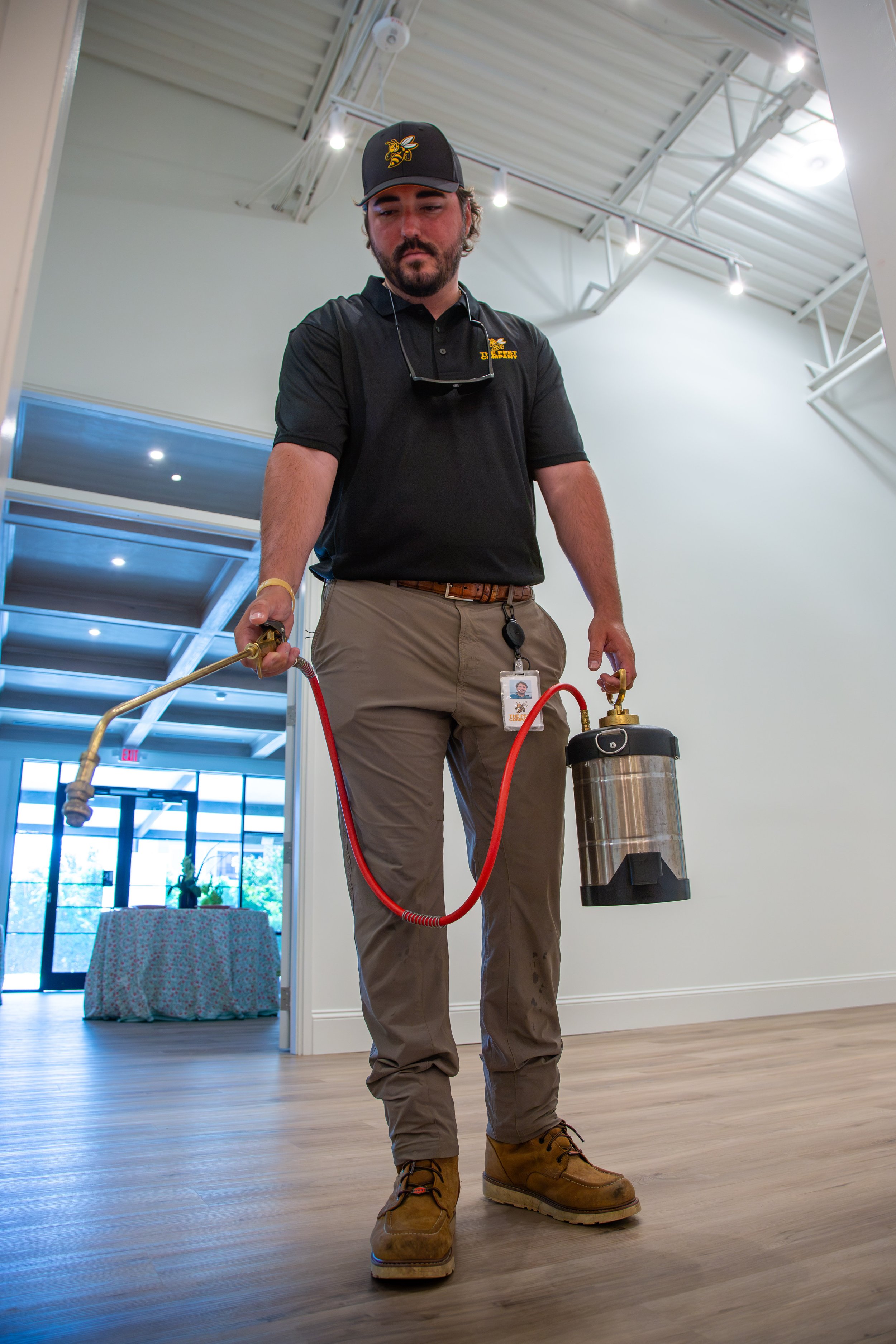 A man wearing a black shirt with a logo, tan pants, and boots is misting a wooden floor with an insecticide sprayer in an indoor space with high ceilings, track lighting, and a large window in the background.
