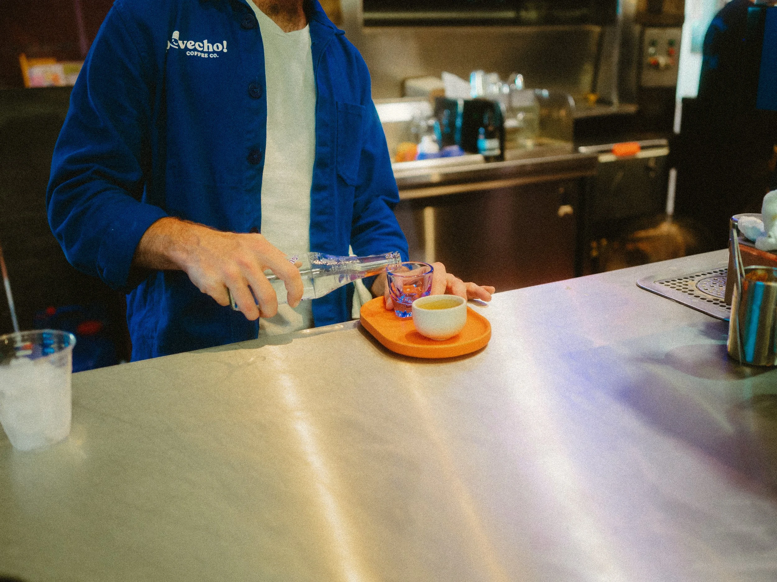 Person pouring a clear drink into a glass on an orange tray, with a small cup beside it, in a kitchen or coffee shop setting.