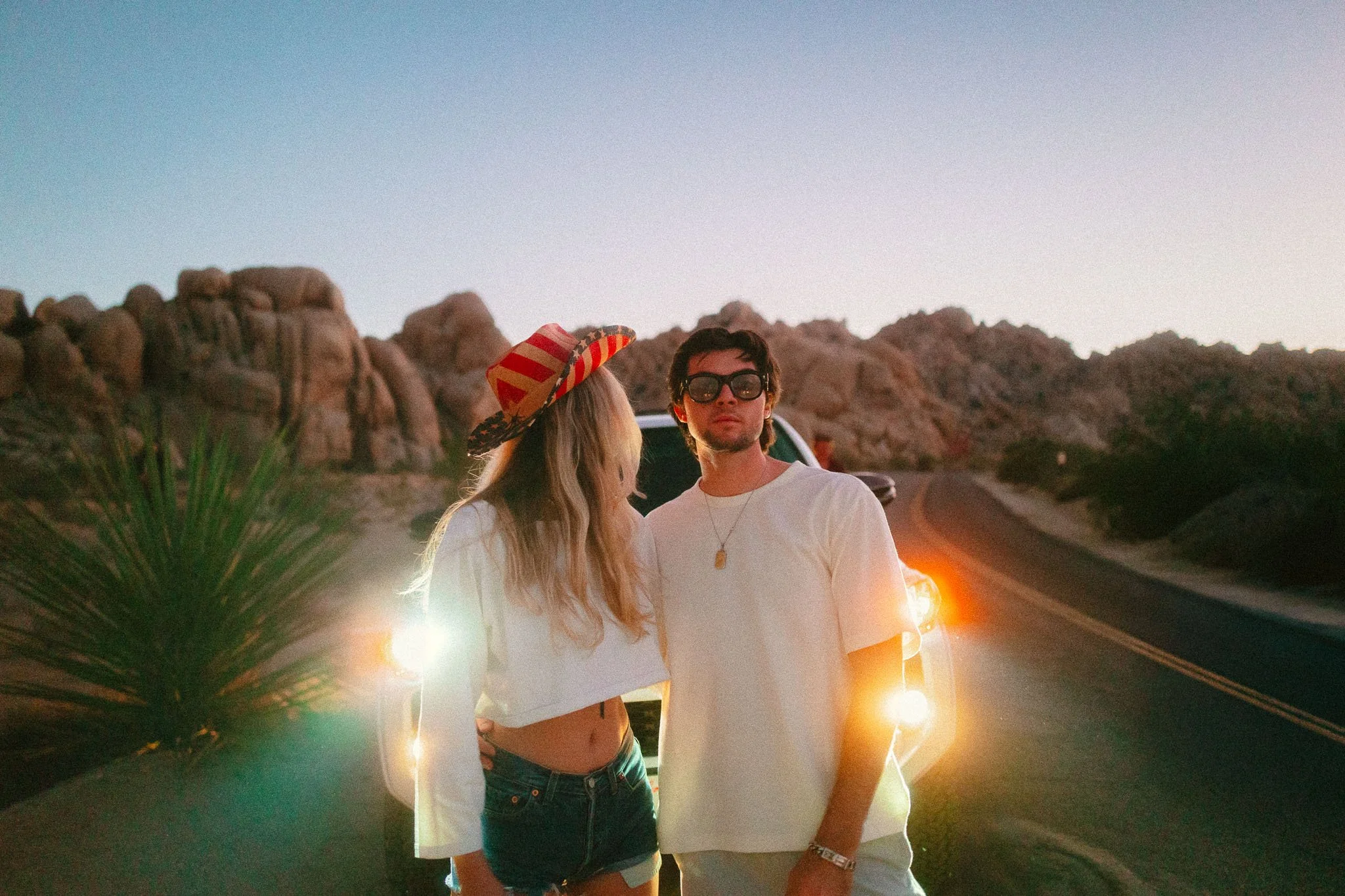 A young man and woman standing on a deserted road at sunset or twilight, with rocky desert landscape in the background. The woman is wearing a large hat and denim shorts, while the man is wearing sunglasses and a white t-shirt, with car lights glowin