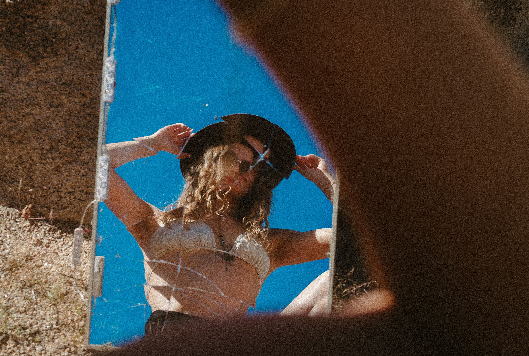 A woman with curly hair wearing a hat and sunglasses is looking at herself in a mirror placed on the ground, with a clear blue sky in the background.