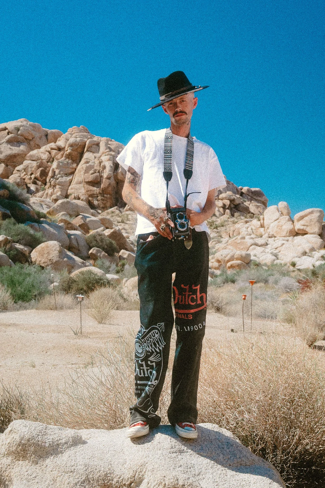 A man with tattoos, wearing a black hat, white t-shirt, and black pants with colorful designs, stands on a large rock in a desert landscape with rocky hills and sparse vegetation.