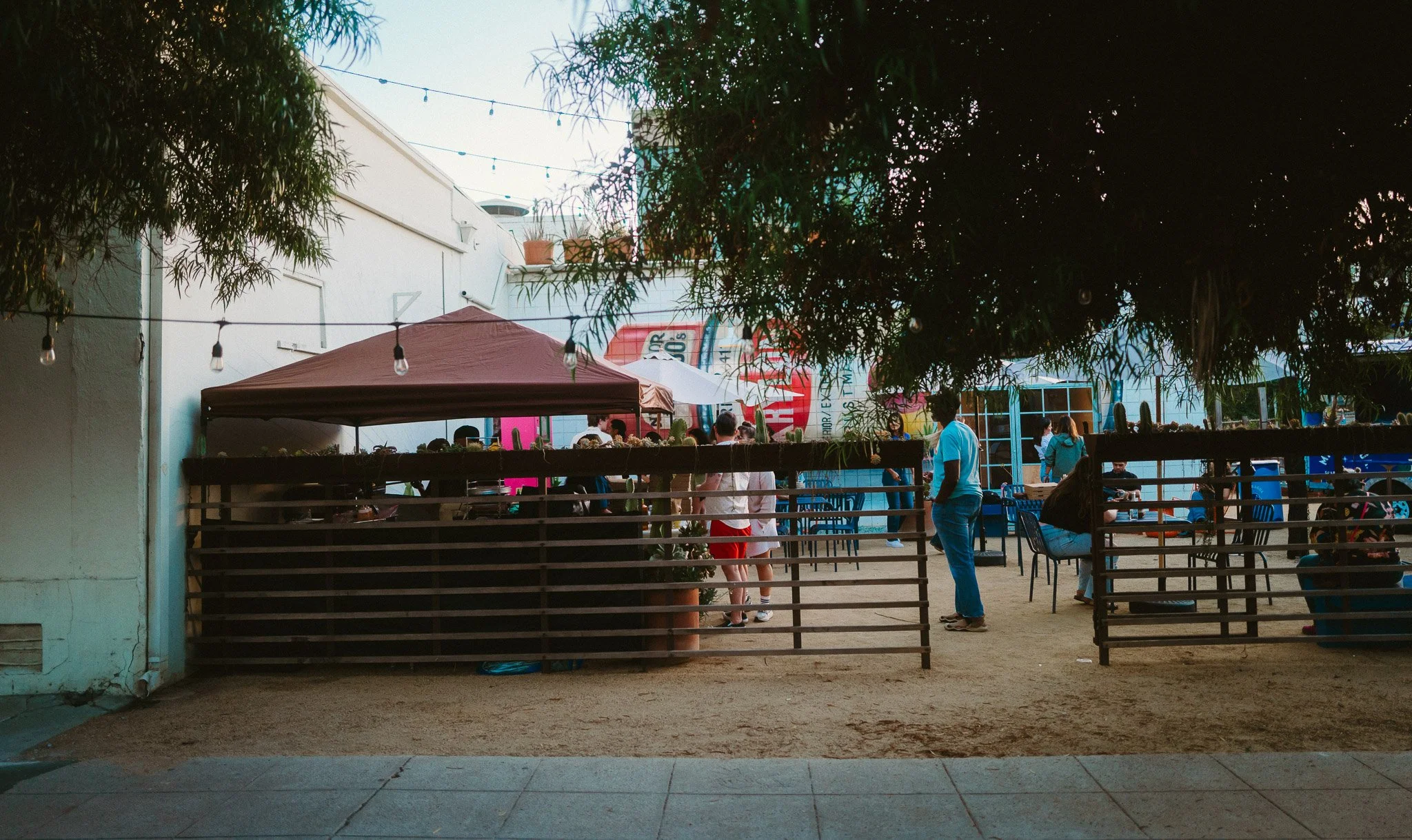 An outdoor dining area with people sitting at tables, some chatting and others eating, surrounded by string lights and trees, with a beige tent and colorful banners in the background.