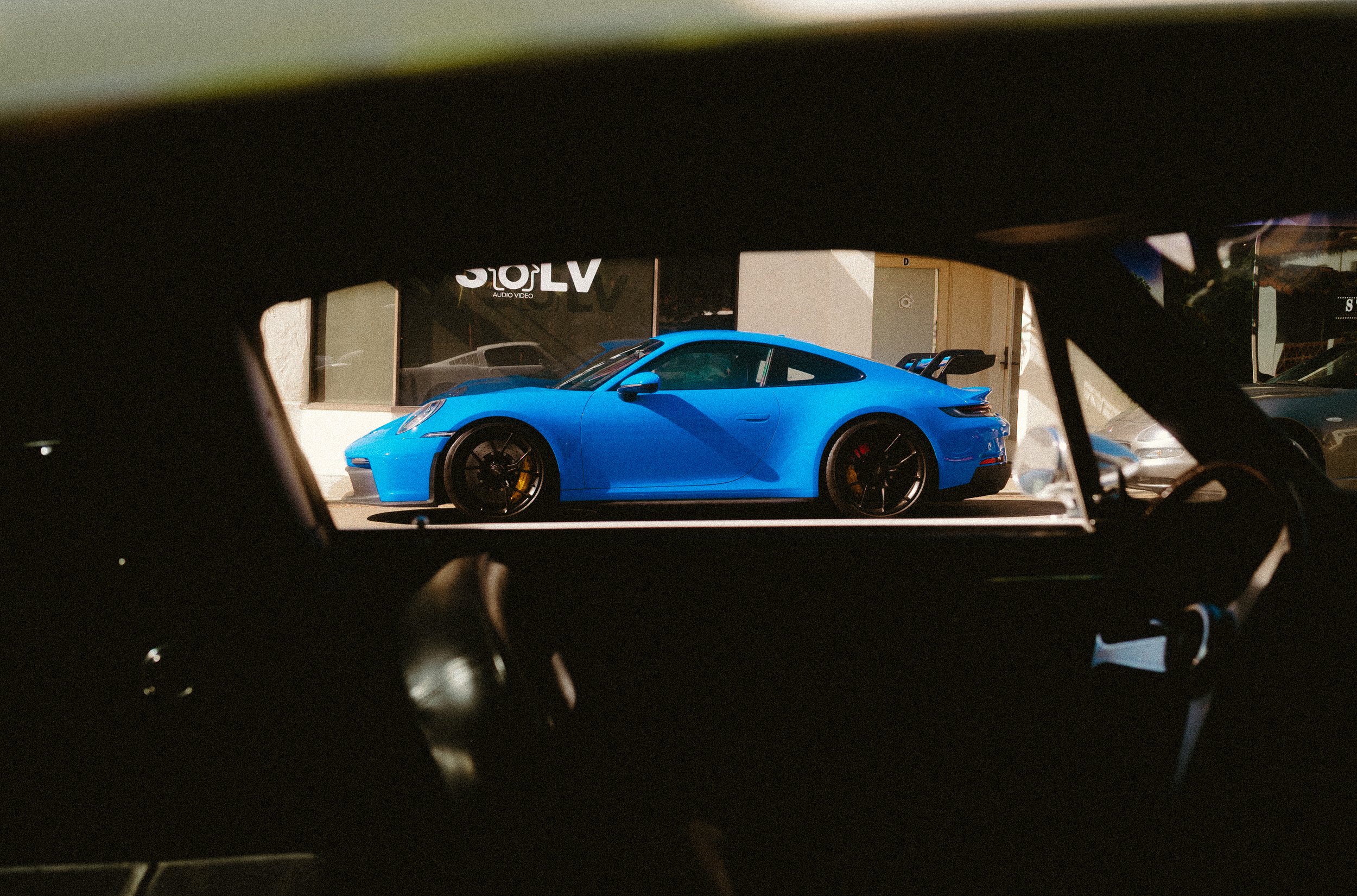 Blue sports car viewed through the windshield and dashboard of a car, parked outside a building.