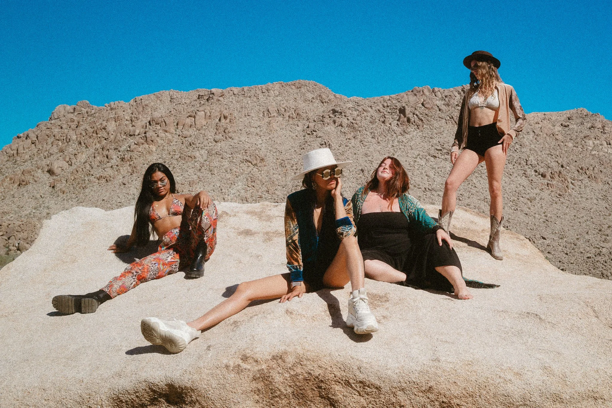 Four women in fashionable outfits sitting and standing on a sandy rock formation in a desert landscape with clear blue sky.