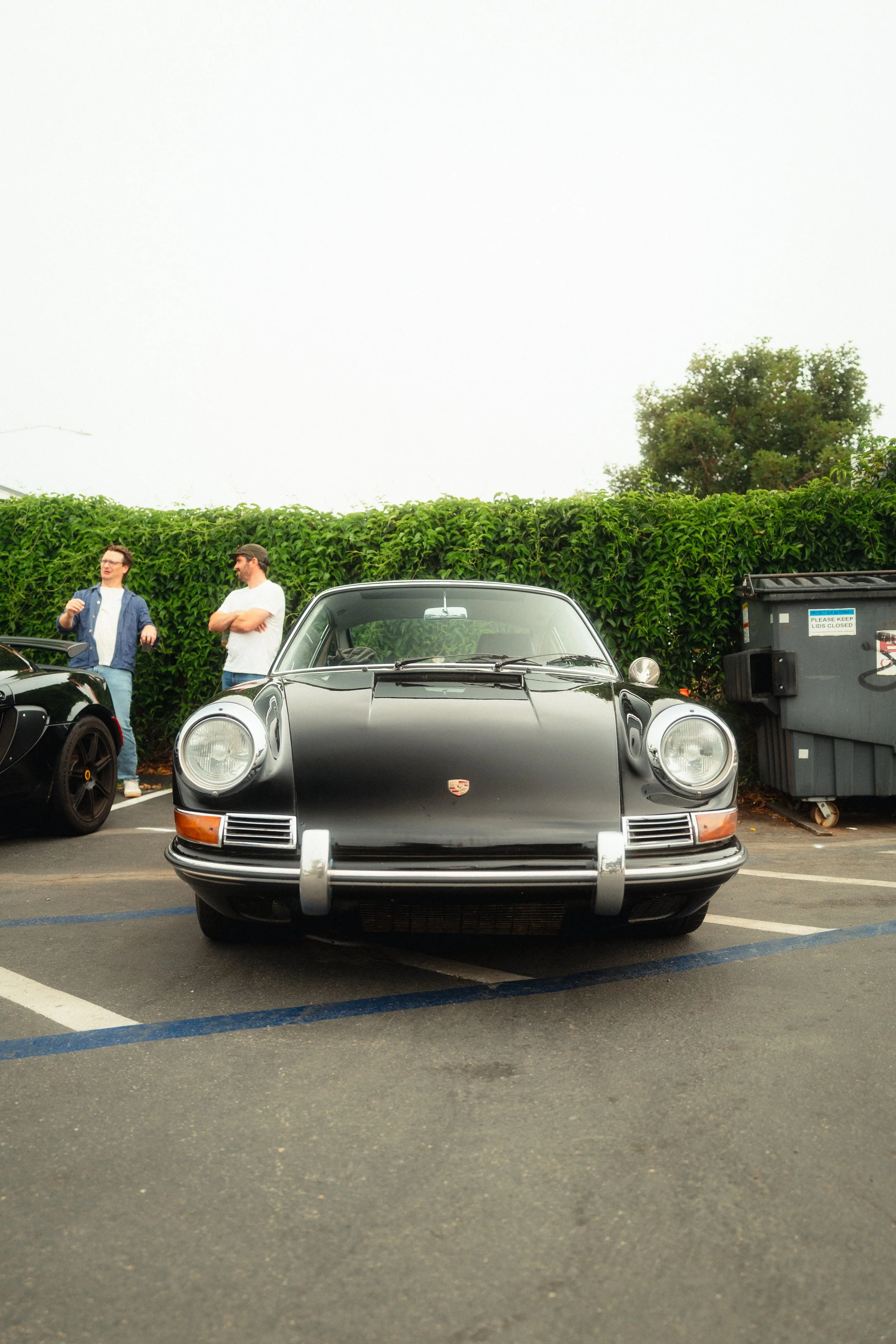 Front view of a vintage black Porsche 911 parked in a parking lot with two men standing and talking in the background, and a green hedge and trash bin behind them.