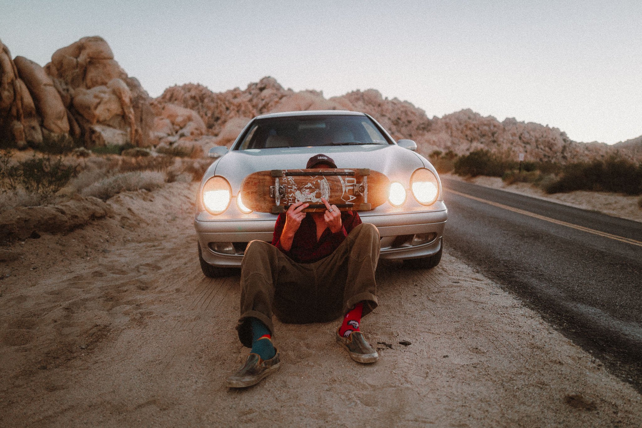 A person sitting on the dirt next to a silver car in a desert landscape, holding a skateboard with a graphic on it in front of their face, with rock formations in the background.