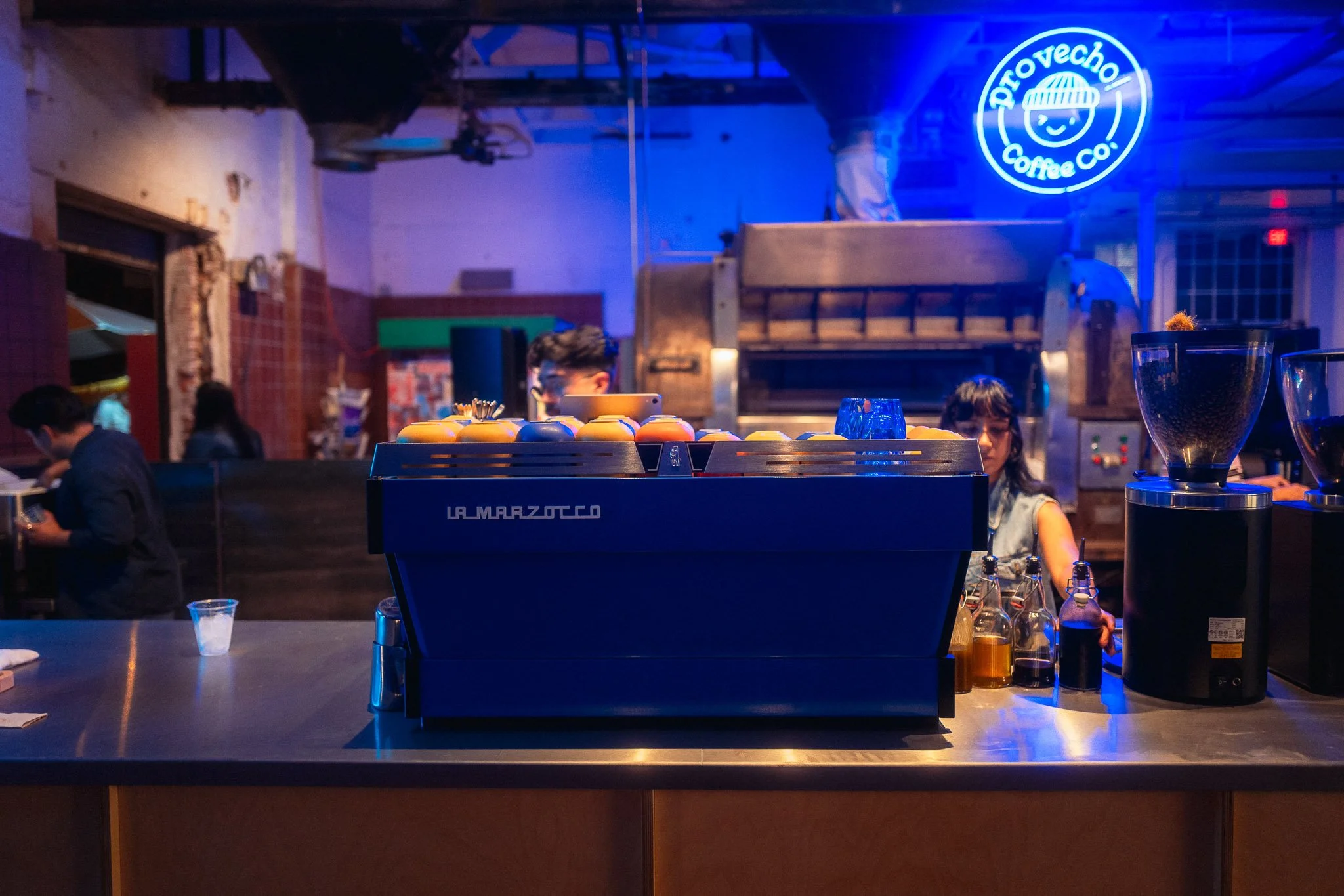 A coffee shop with a La Marzocco espresso machine on the counter, bottles of syrups, and a grinder. Background includes a neon sign for ProtoVeco Coffee Co., and workers preparing coffee in a dimly lit, industrial-style interior.