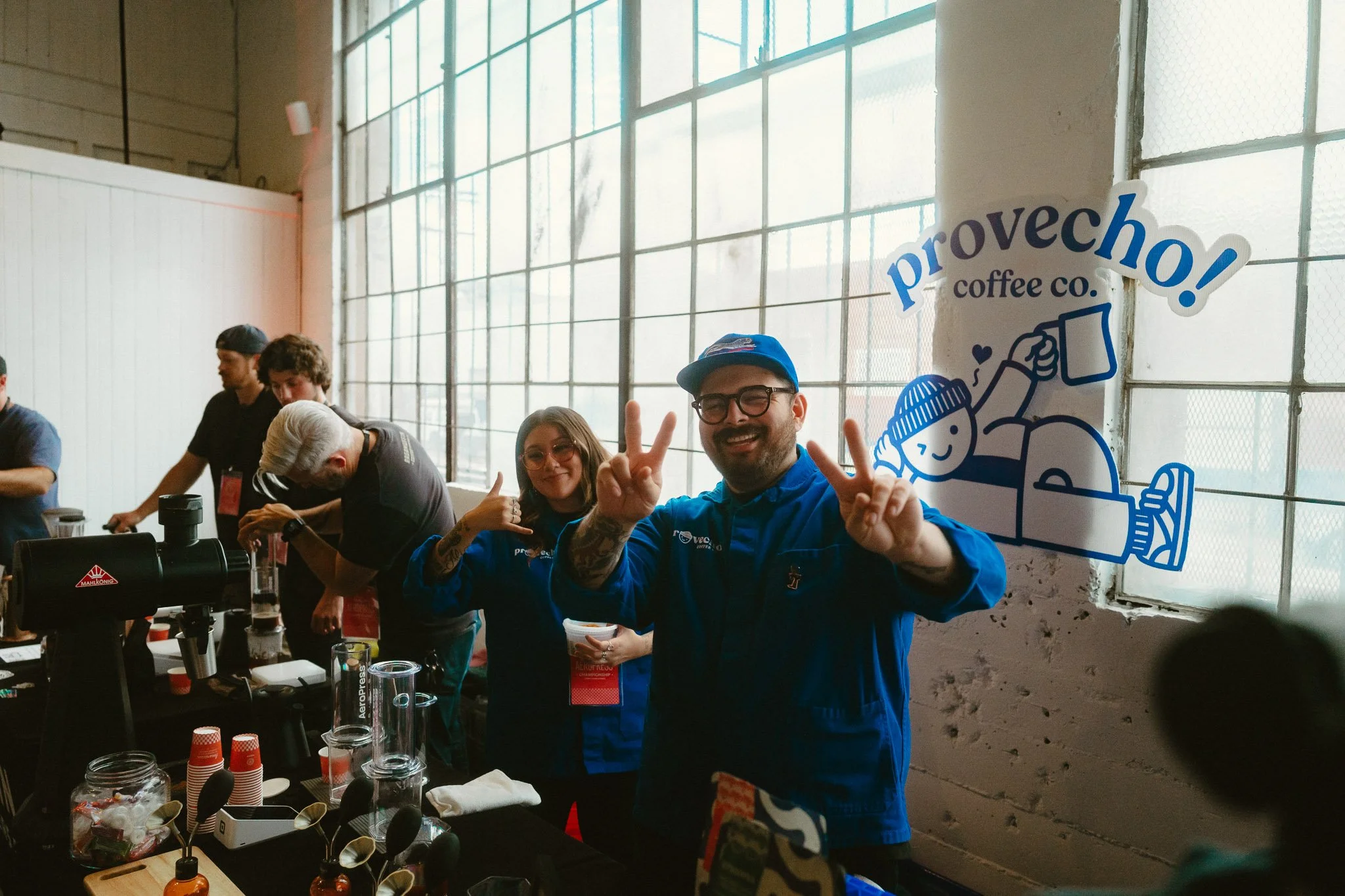 Group of people working behind a coffee bar at a coffee shop event, with two smiling individuals in the foreground making peace signs, in front of a wall with a fun sign that says 'proveccho! coffee co.' with a cartoon illustration of a person holdin