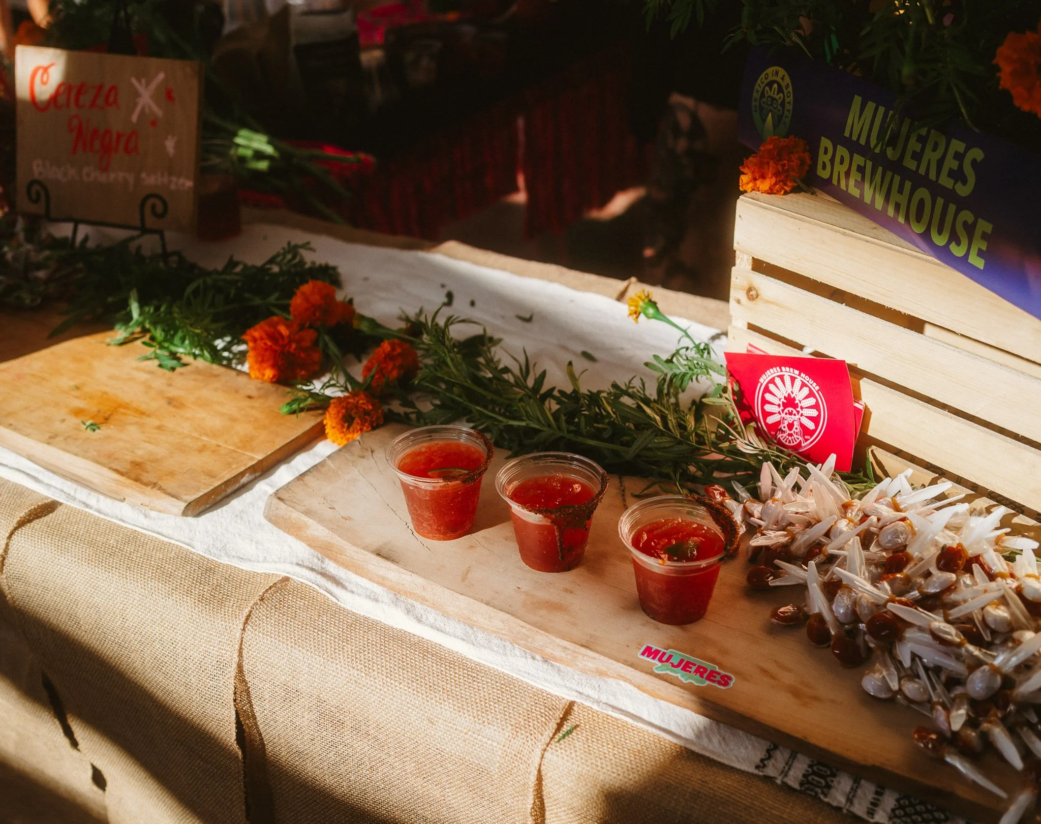 A display of small cups with red sauce and chilies, marigold flowers, and signs indicating 'Mujeres Brewhouse' and 'Cerveza Negra' at a market stall.