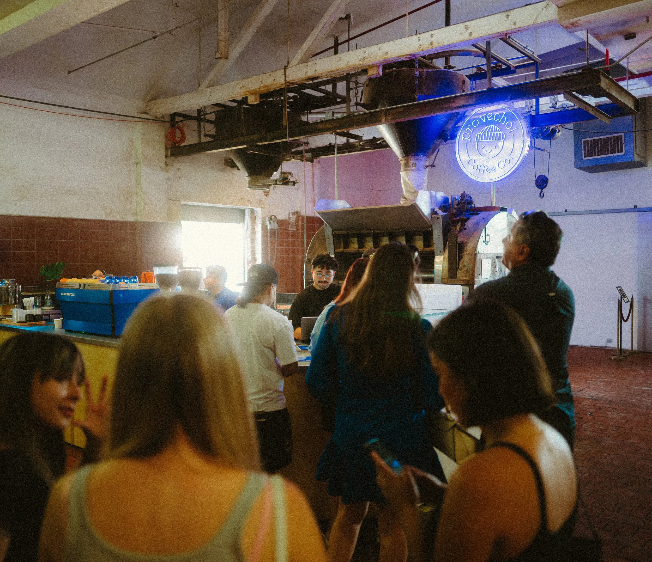 People standing and sitting inside a coffee shop with a large coffee roasting machine visible in the background, illuminated by a neon sign that says 'Provecho Coffee Co.'
