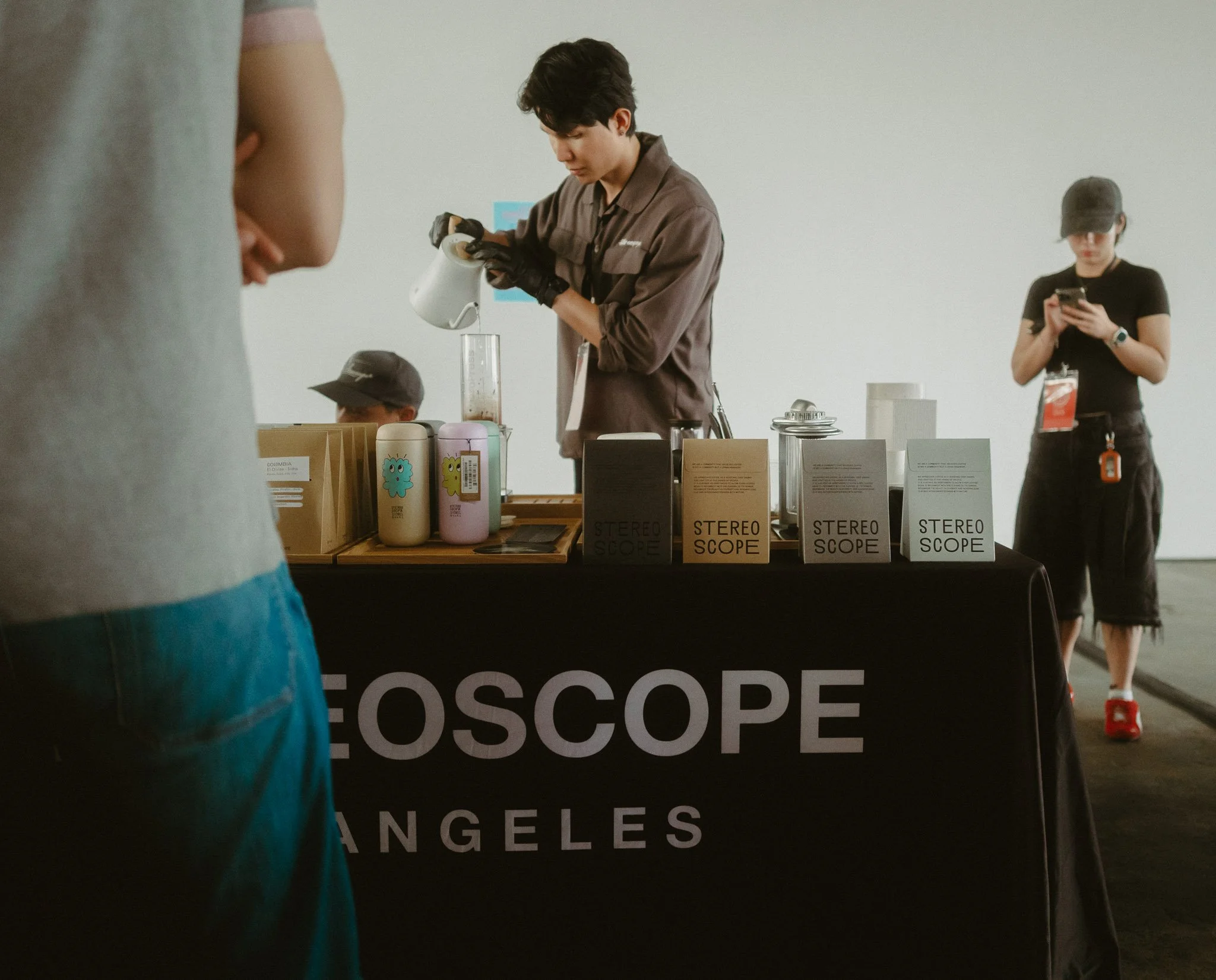 Person preparing a drink behind a table with boxes labeled "STEREO SCOPE" and colorful containers, while a woman in black checks her phone in the background at an indoor event.