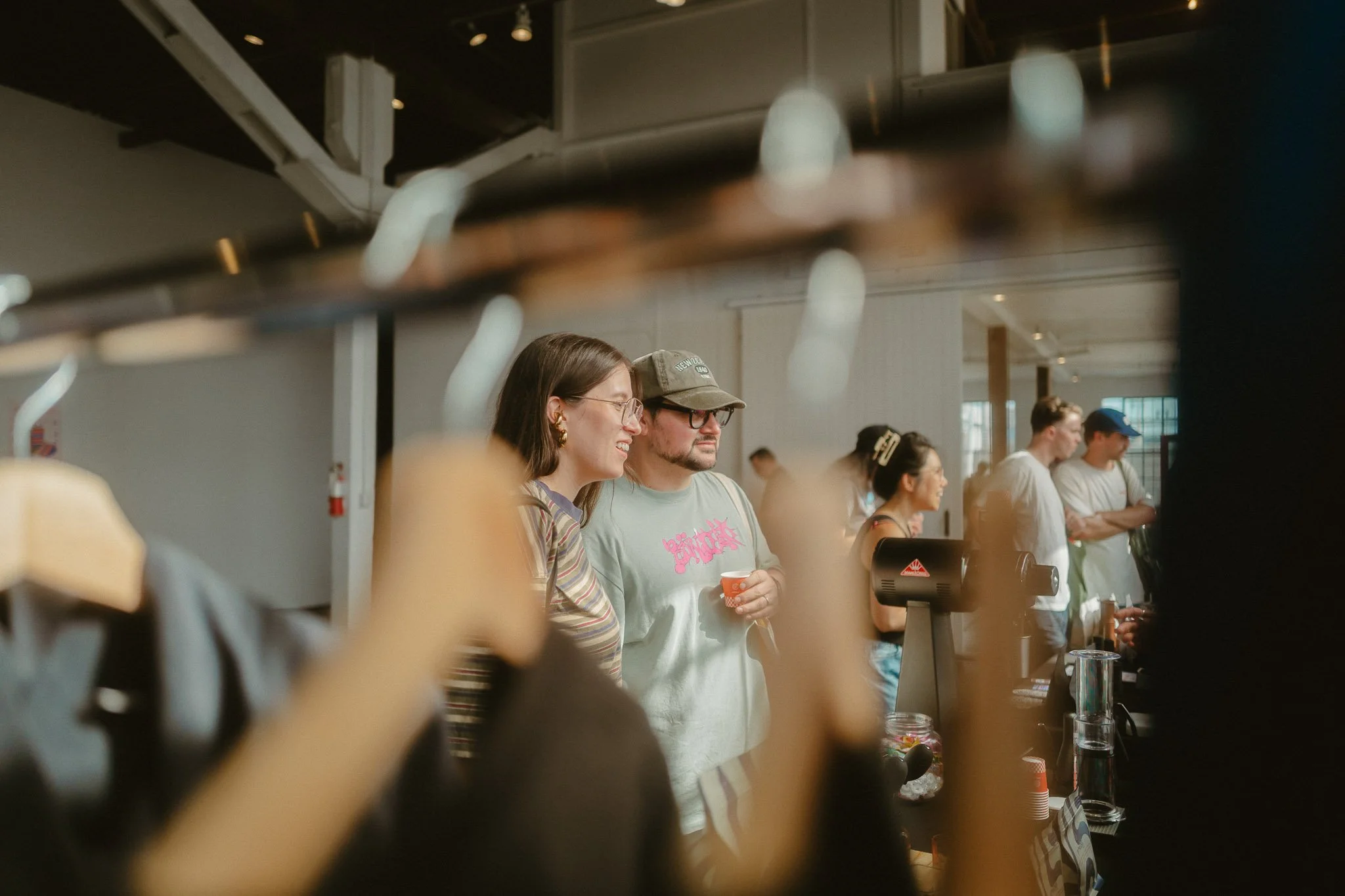 People gathered at a bar or café, seen through a blurry object in the foreground, with some individuals engaging in conversation and others waiting.