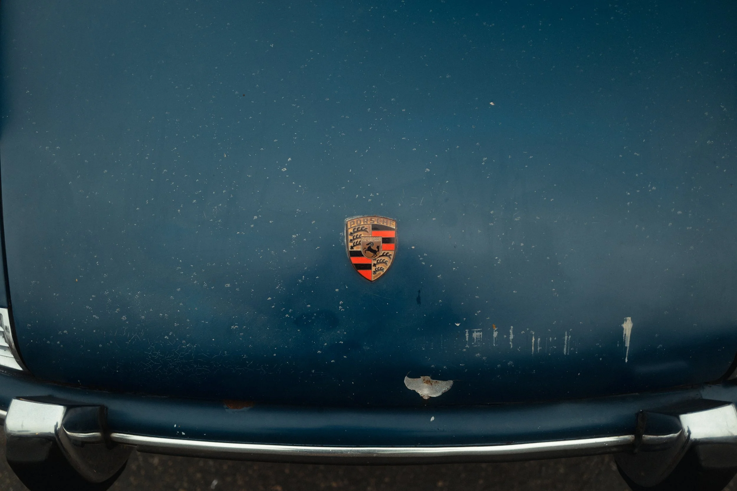 Close-up of the hood of a blue Porsche car with a rusted and chipped paint and the Porsche emblem in the center.