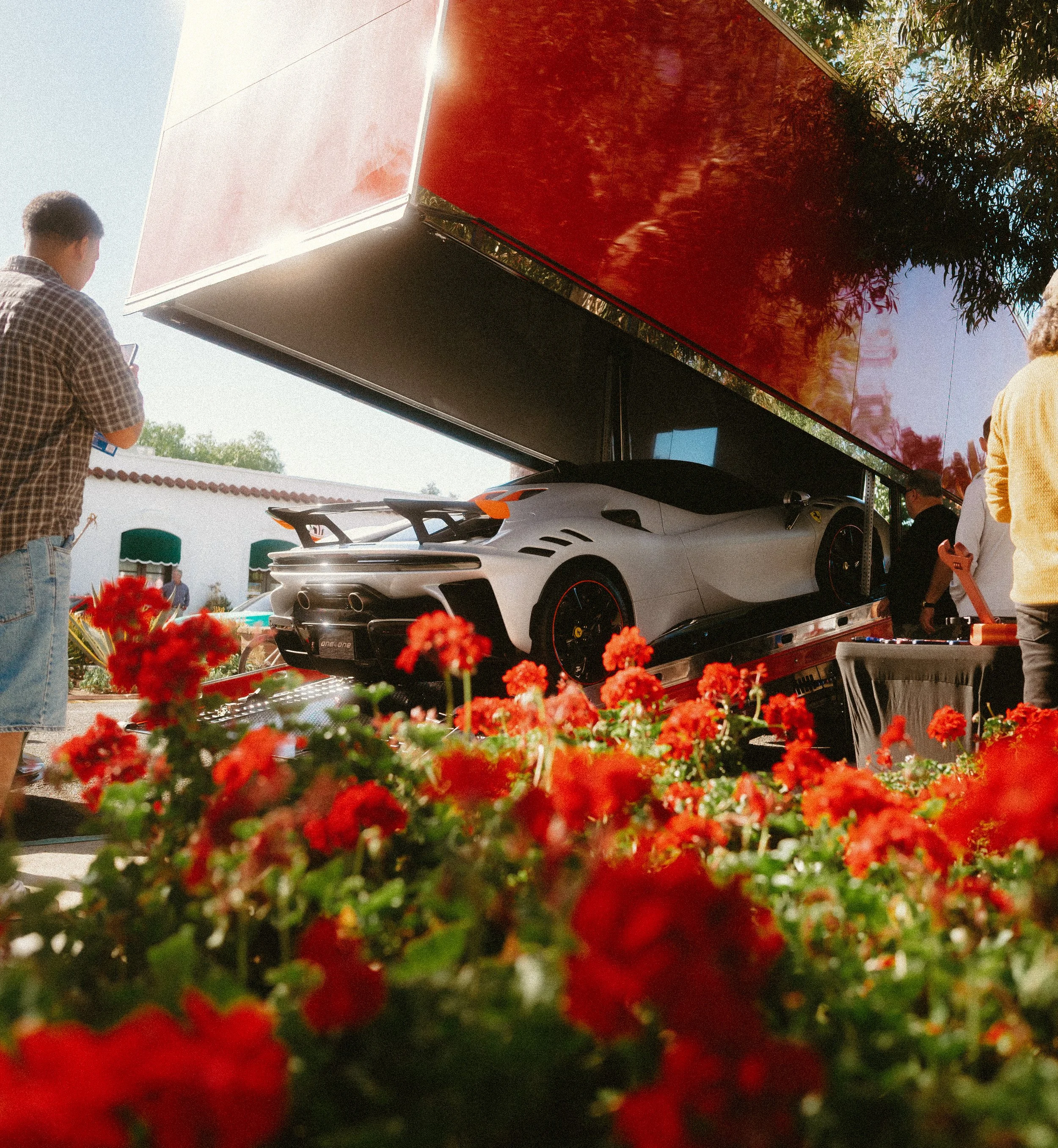 A sleek, white sports car displayed on a platform under a large red canopy, with people around it at an outdoor event and red flowers in the foreground.