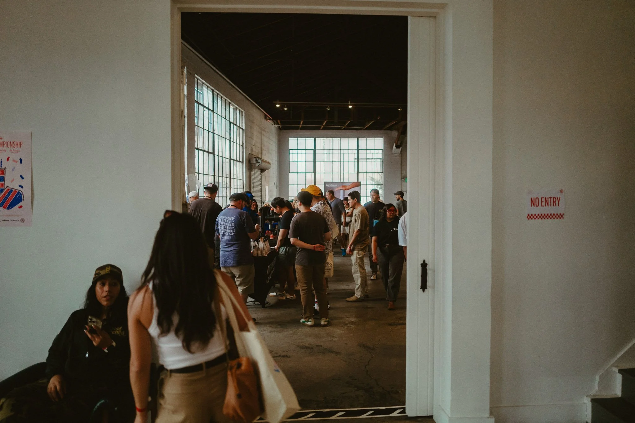 Scene of people gathered inside an industrial-style building with large windows, some standing in line or browsing at a booth.