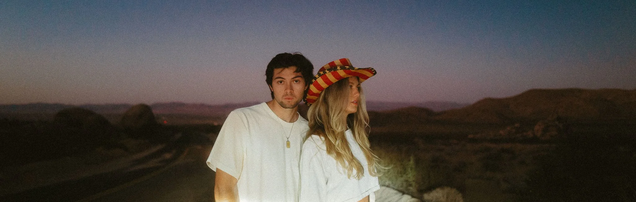 A young man and woman standing outdoors at sunset, with mountainous landscape in background. The woman is wearing a large, colorful striped hat, and both are dressed in white shirts.