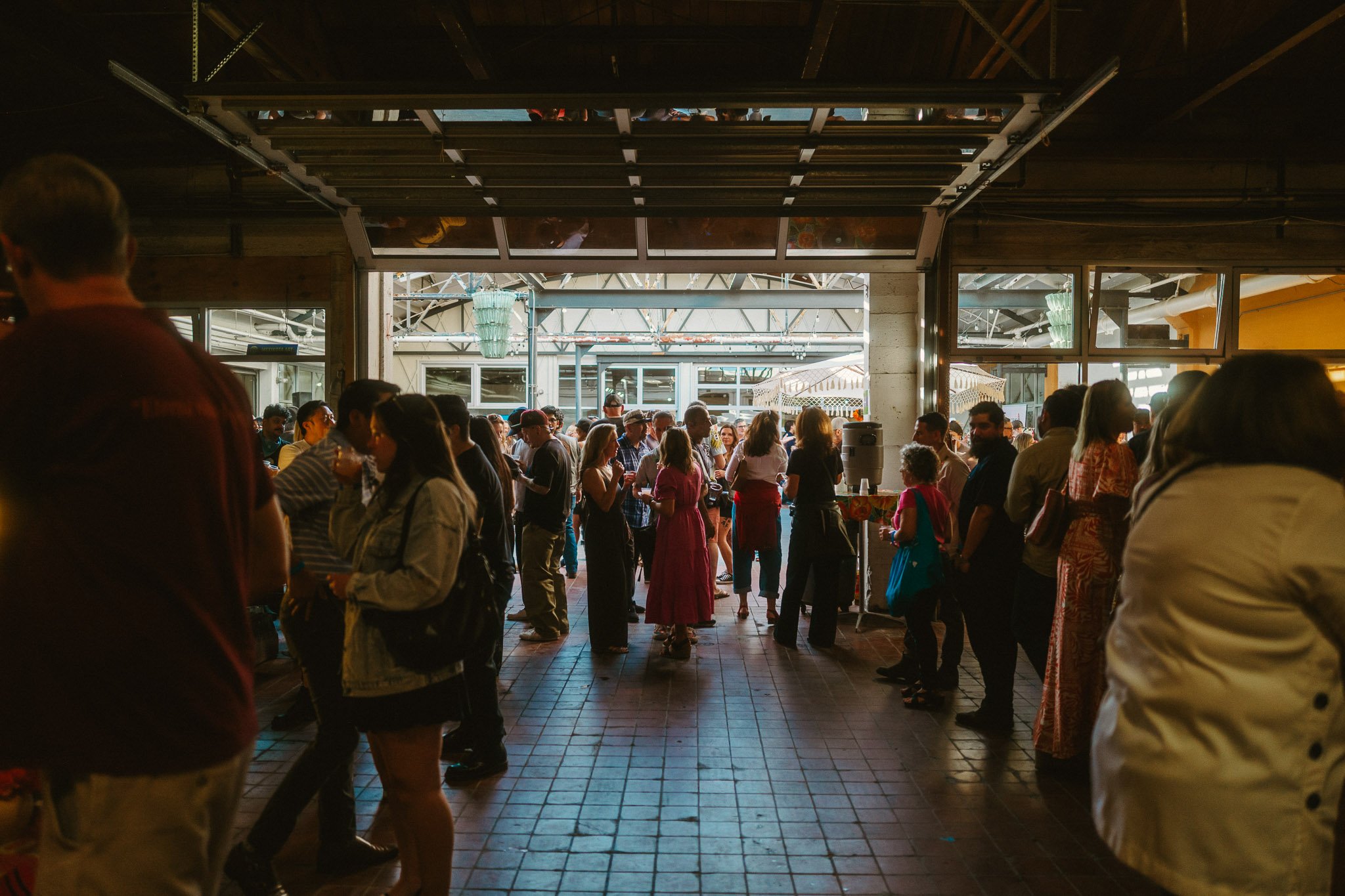 Crowd of people gathered inside a building near an open garage door, illuminated by natural light from outside, with some people standing and conversing, and others walking.