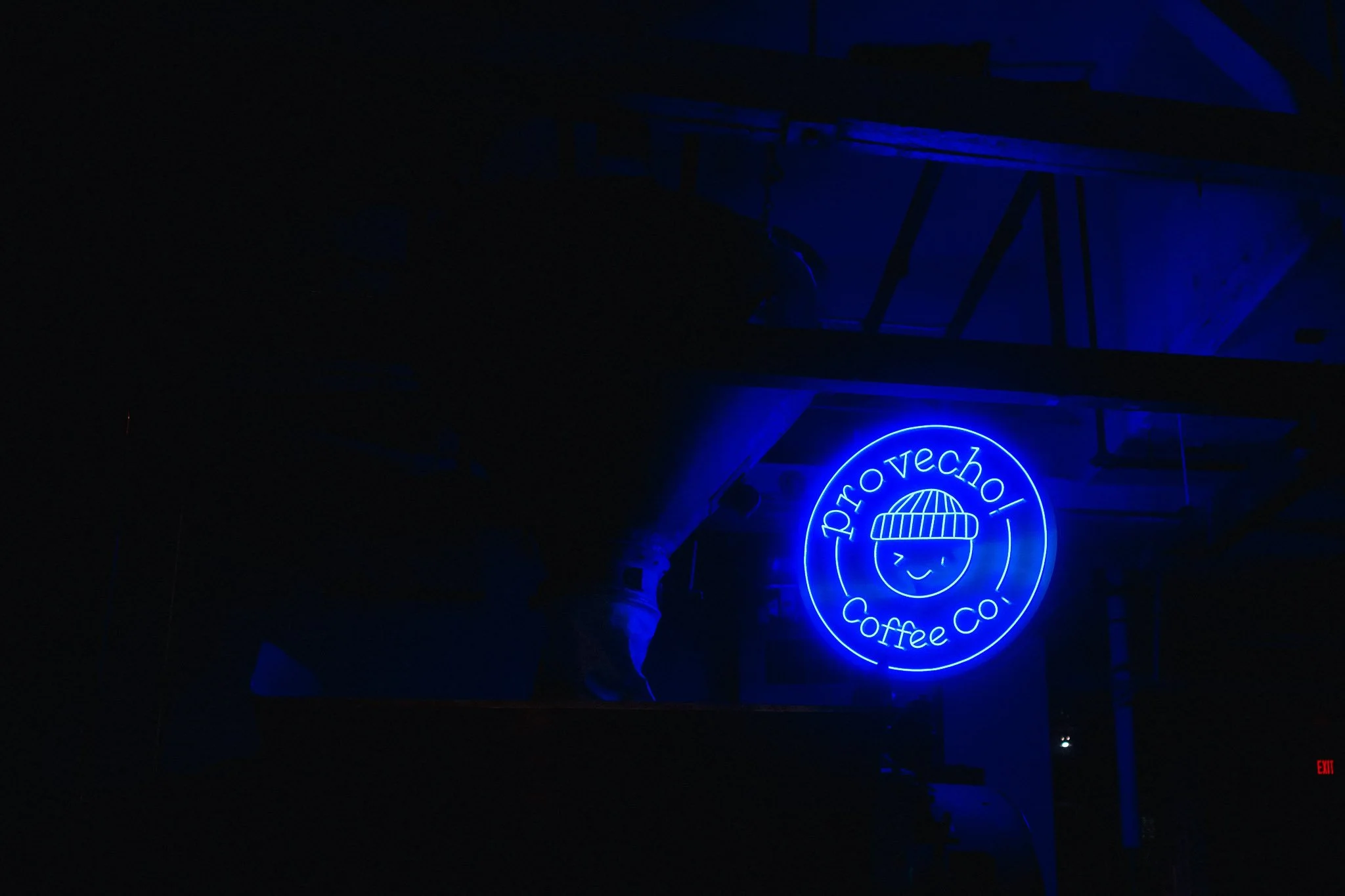 Blue neon sign for Pro Vechi Coffee Co. with a smiling face wearing a beanie, inside a circle, in a dark room.