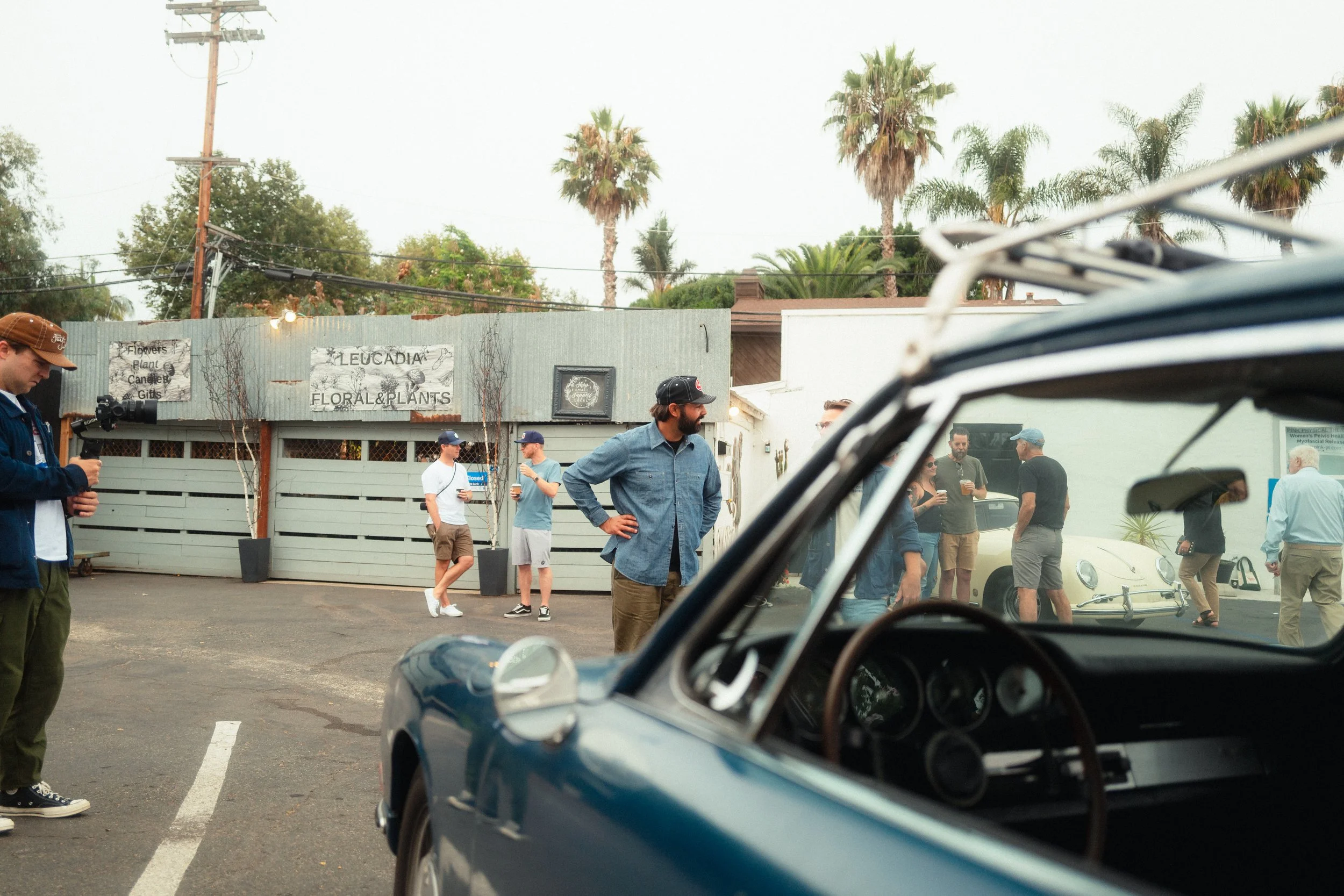 People gather outside a floral shop, Leucadia Floral & Plants, with some holding drinks and conversing, as a classic blue car is partially visible in the foreground.