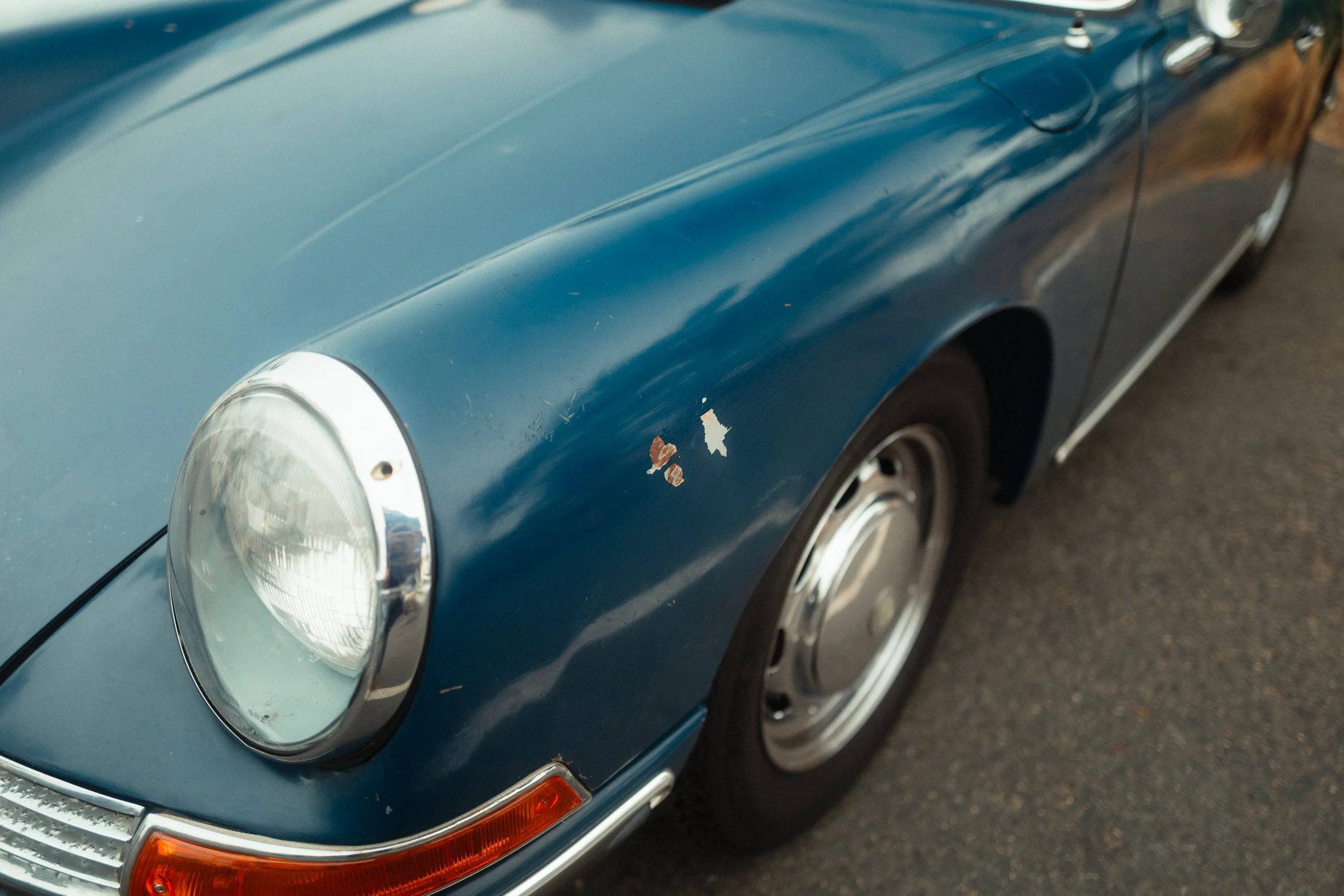 Close-up of the front left part of a vintage blue sports car with a round headlight, chrome details, and a wheel. The car has some paint chips and rust spots.