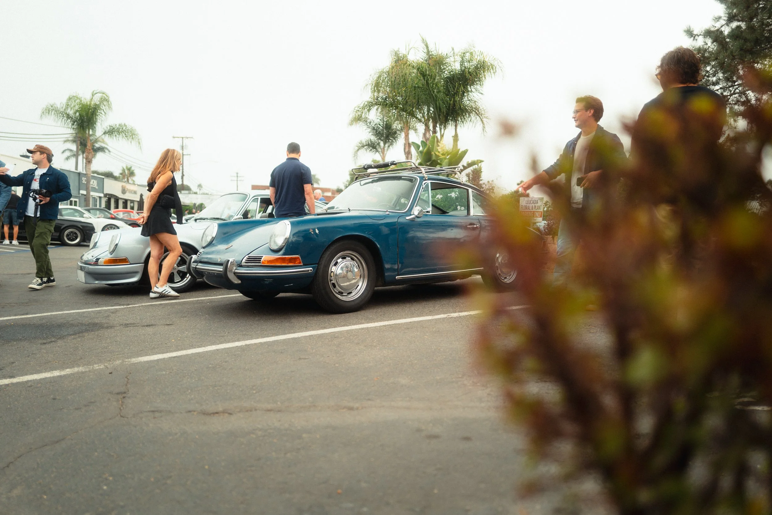 People gathered around classic cars parked in a lot, with palm trees and shops in the background, during daytime.